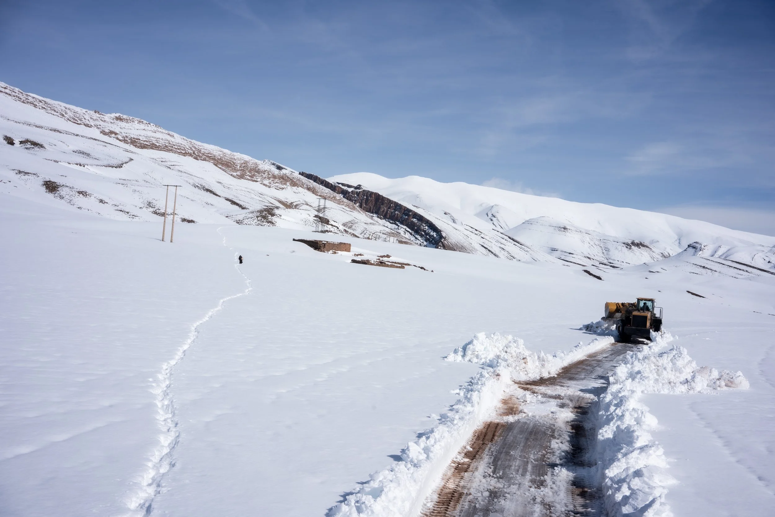 Two distinct lifelines across the landscape: the freshly cleared asphalt carved by machinery and a lone trail of footprints leading up the frozen slope. In the high terrain, every cleared meter and hand-traced path marks a return to connection.