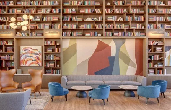 A cozy lounge area with a large wooden bookshelf filled with books, a long gray sofa, colorful armchairs, small round coffee tables, abstract artwork on the wall, and modern lighting fixtures.