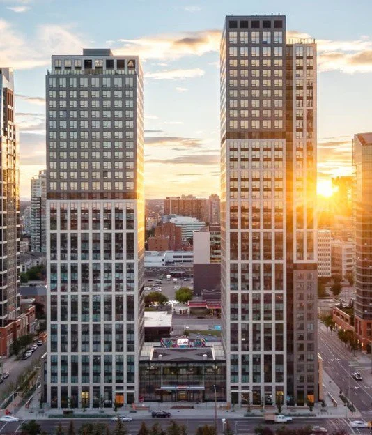 Two tall modern skyscrapers with glass facades in a city at sunset, with the sun partially visible on the right side.