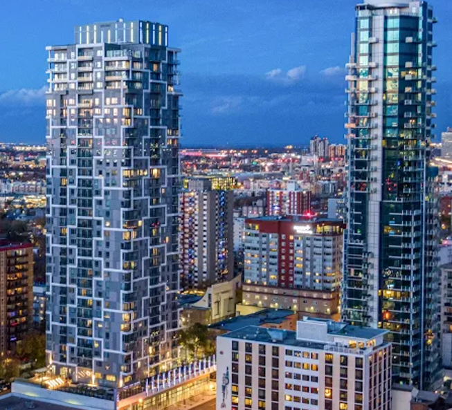 City skyline at dusk featuring two modern high-rise buildings with glass facades, surrounded by other city buildings and lights.