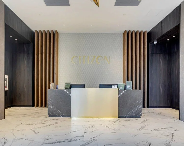 Modern reception area with a white marble desk, dark marble accents, and wooden slats on the wall. The wall behind the desk has a large 'CITIZEN' sign in gold letters.