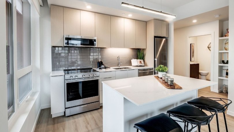 Modern kitchen with white cabinets, gray backsplash, stainless steel stove and microwave, white island with black barstools, and a view of a bathroom in the background.