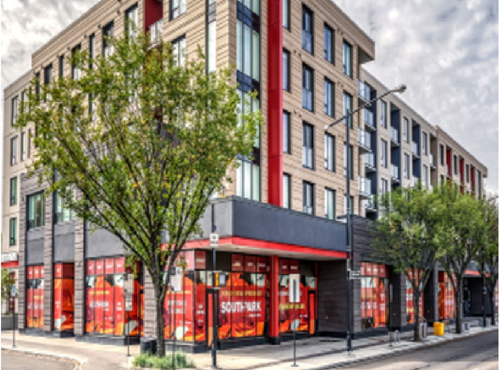 A multi-story building with retail stores on the ground floor, featuring large windows and red accents. There are trees along the sidewalk, with some greenery visible. The building appears to be in an urban area with a street, streetlights, and a cloudy sky.