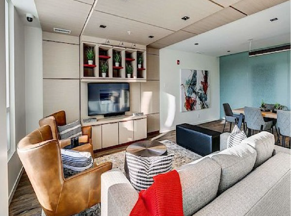 Modern living room with a beige sofa and a pair of brown leather armchairs. A flat-screen TV is mounted on a built-in wall unit with decorative plants on shelves. There's an abstract painting on the wall and a dining area with chairs and a table in the background.