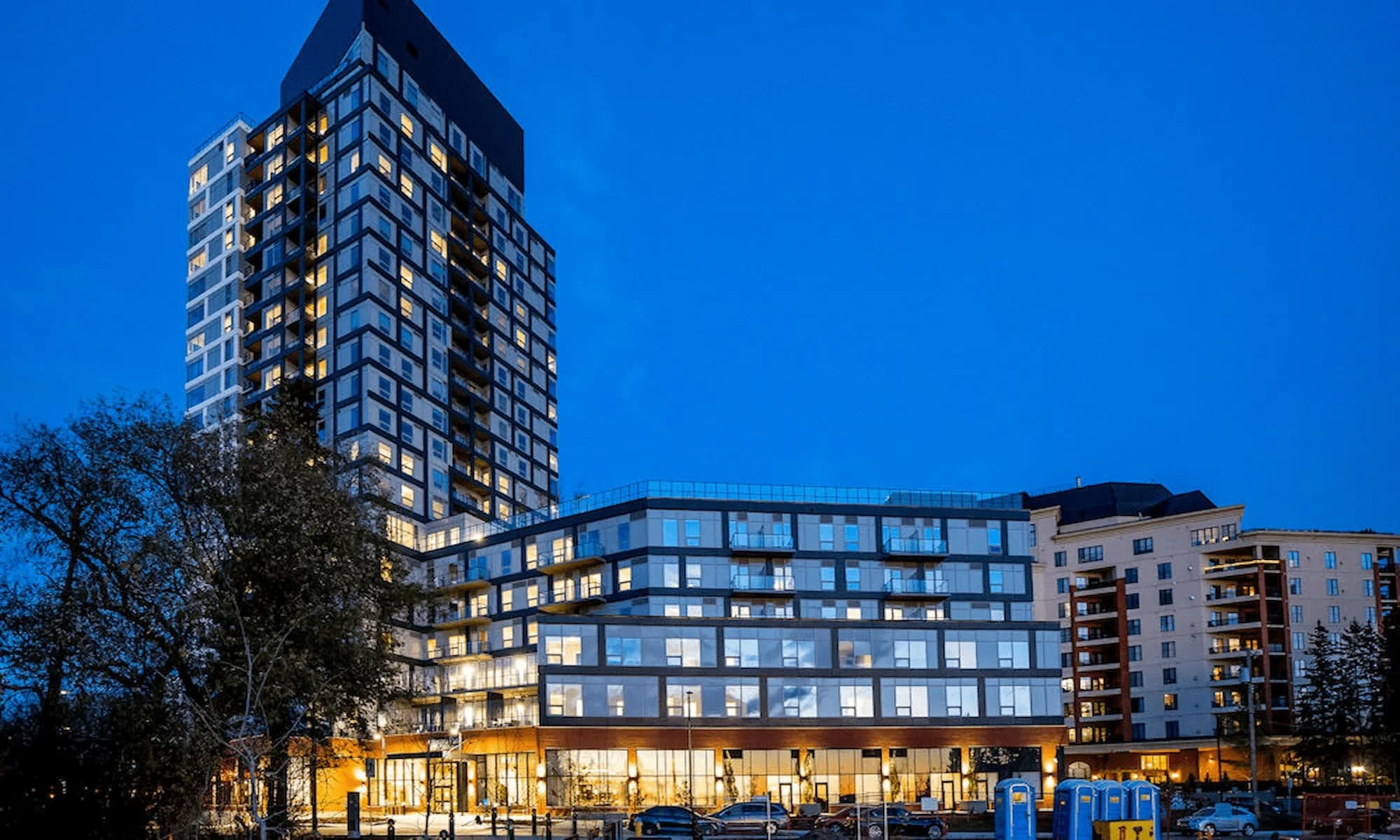 A modern multi-story apartment building at dusk with illuminated windows and a parking lot in the foreground.