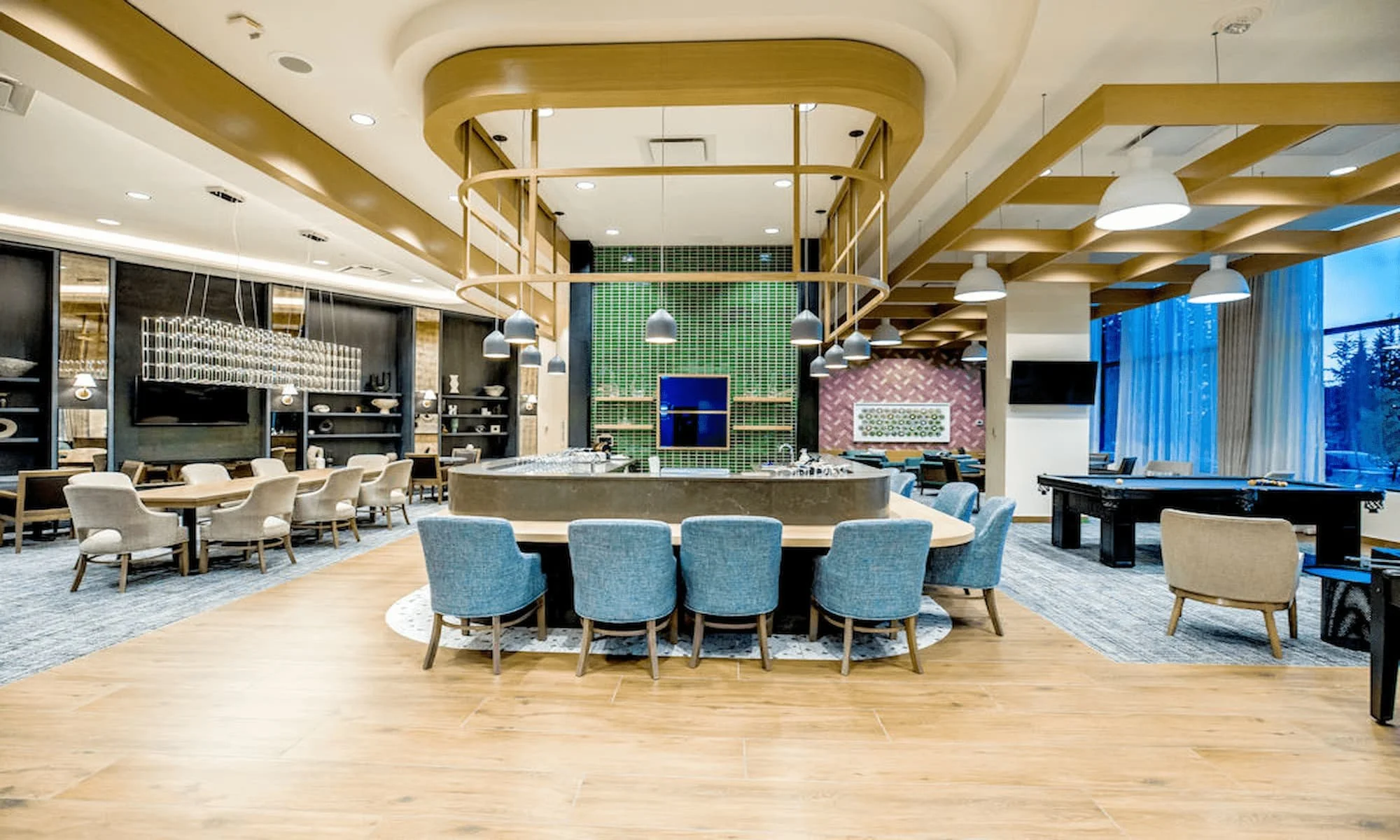 A spacious, modern lounge area with a large central bar surrounded by blue chairs, a black grand piano on the right, and various tables and chairs. The room features wood accents, a high ceiling with pendant lights, and large windows with blue curtains.