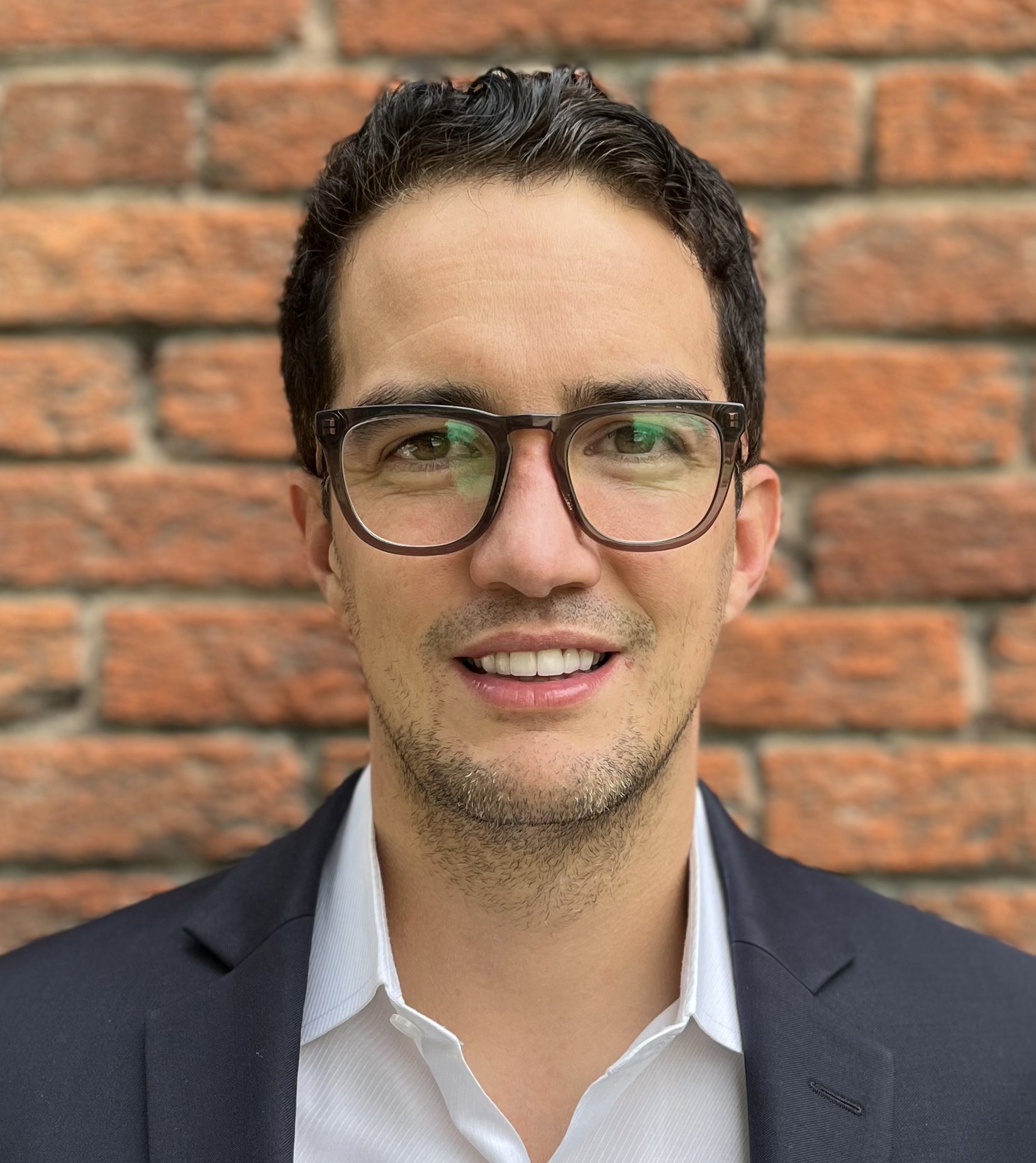 A man with dark curly hair, wearing glasses, a white shirt, and a dark blazer, smiles in front of a brick wall.