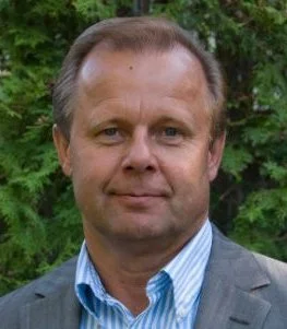 A man in a gray suit and blue striped shirt standing outdoors with green trees in the background.