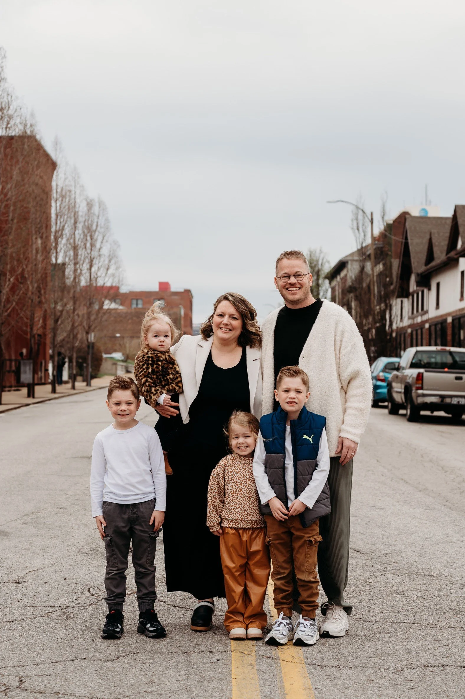 A group of six people, including adults and children, standing on a street. The adults are smiling and the children are standing in front. The background shows residential buildings and parked cars, with leafless trees and an overcast sky.