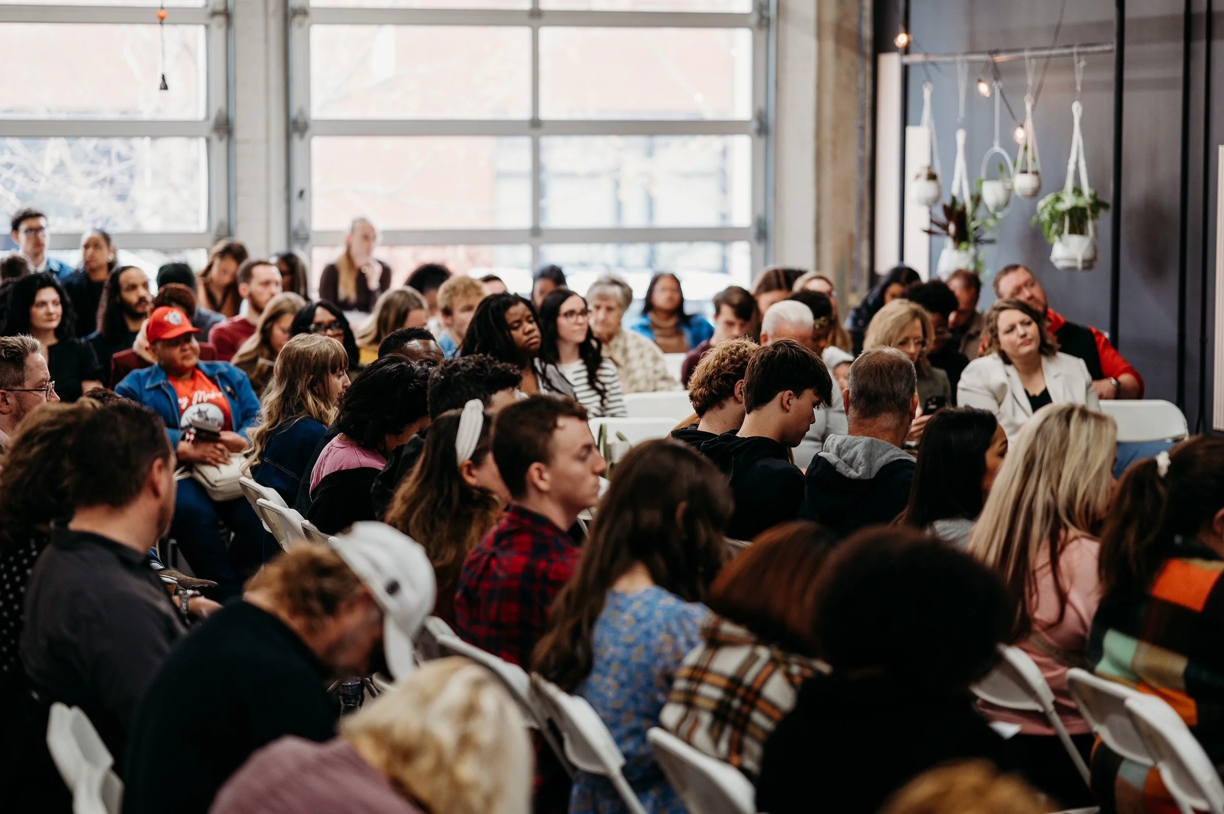 A large group of people sitting on white chairs inside a modern building with large windows and hanging potted plants, attending a conference or seminar.