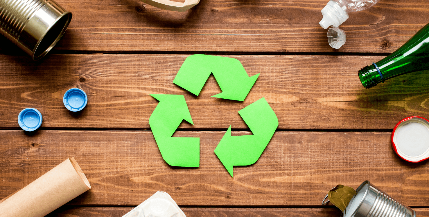 Recycling symbol made of green paper on wooden surface surrounded by various recyclables such as aluminum cans, plastic bottles, bottle caps, and a cardboard tube.