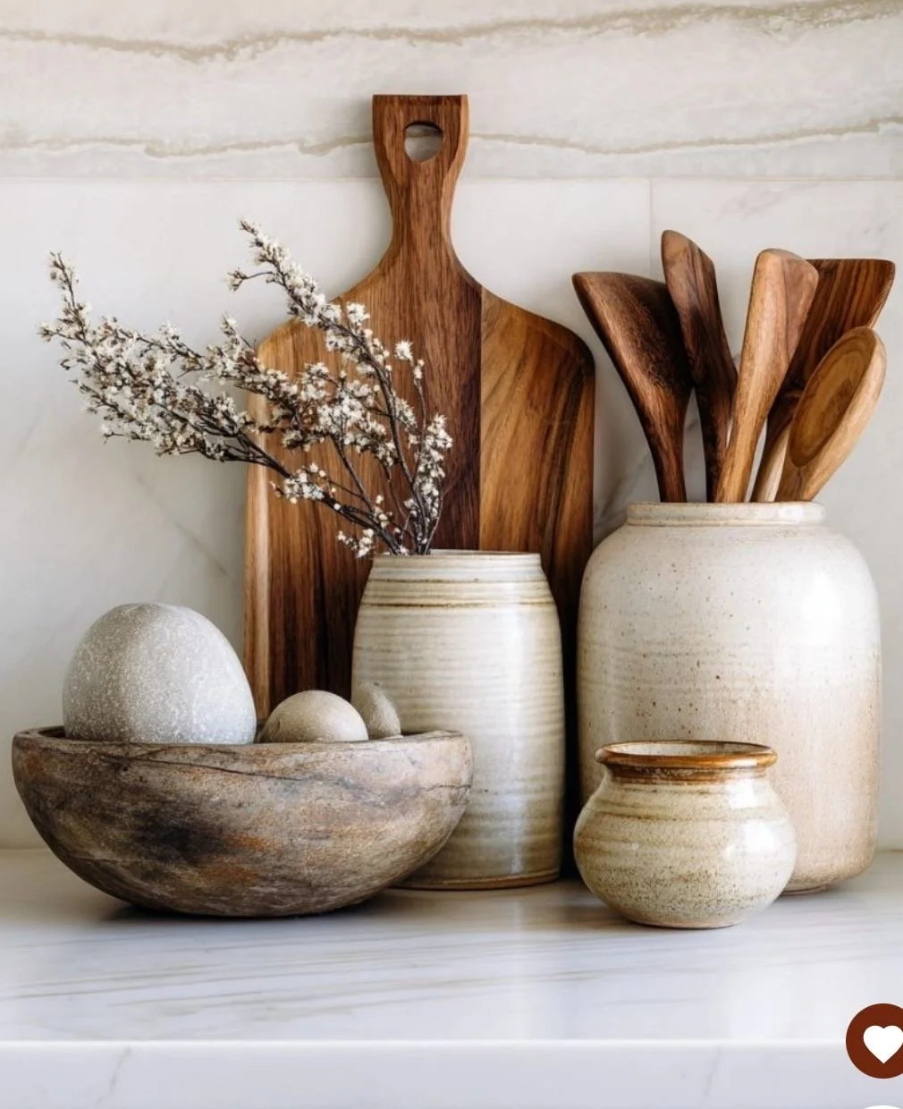 Decorative arrangement of wooden kitchen utensils in ceramic jars, a wooden cutting board, and a bowl with decorative eggs and branches on a marble countertop.