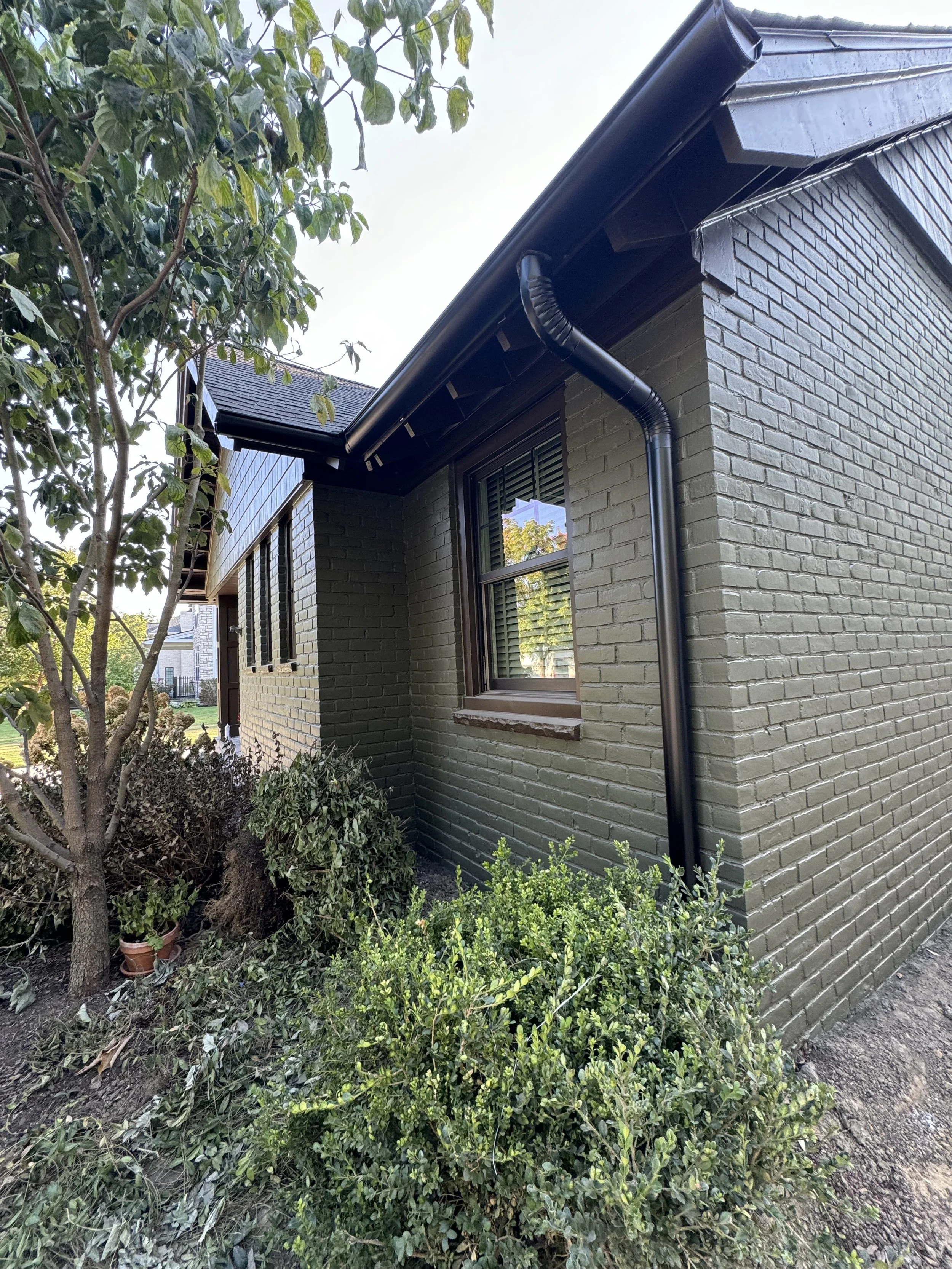 Side of a house with dark green brick exterior, double pane window, black rain gutter, surrounded by bushes and a tree.seamless gutters installed by dads seamless gutters in michigan