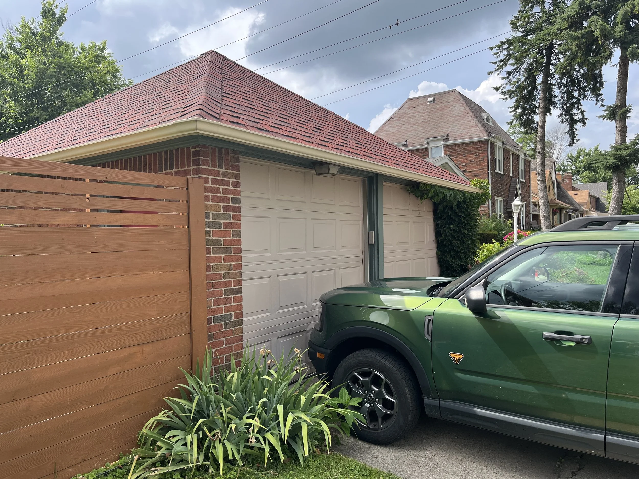 A residential driveway scene showing a green Range Rover parked in front of a garage with a white door and a brick and wood fence. dads seamless gutters cleaned there gutters
