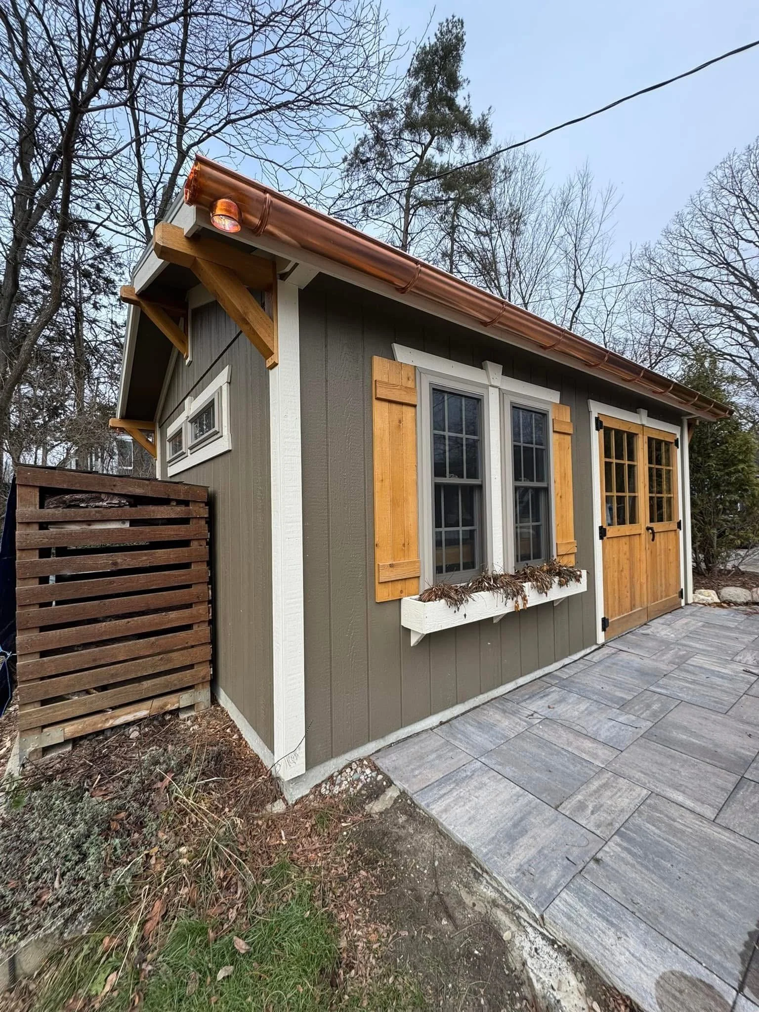 Small backyard shed with gray vertical siding, wooden trim, and a copper-gutters installed by dads seamless gutters in Milford michigan, It has double-hung windows with wooden shutters and a sliding barn door, with a paved outdoor area in front.