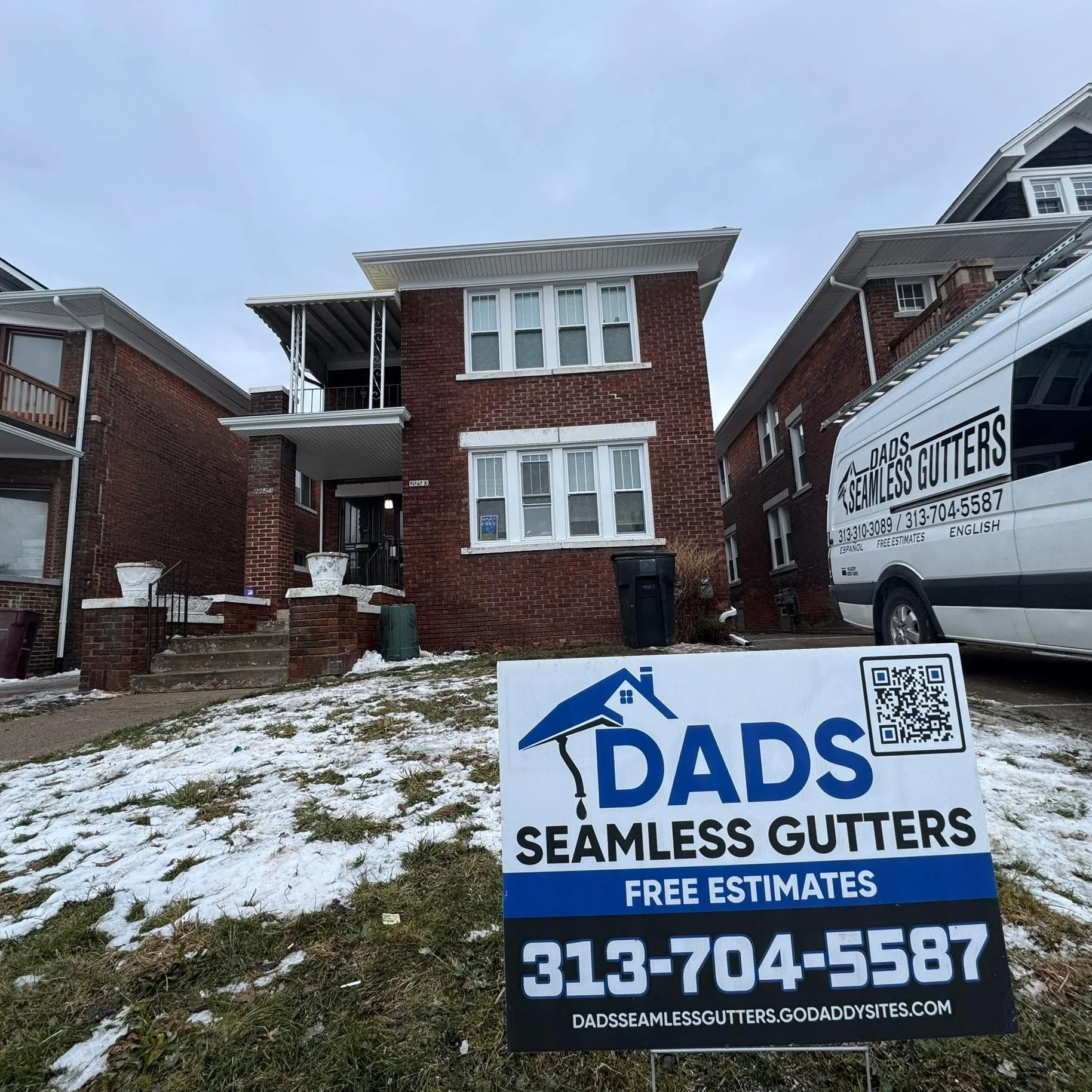 Front view of a red brick duplex house with a small front yard, steps leading up to the entrance, and a sign for Dads Seamless Gutters on the lawn. A van with the gutter company's branding is parked beside the house, and snow is on the ground.