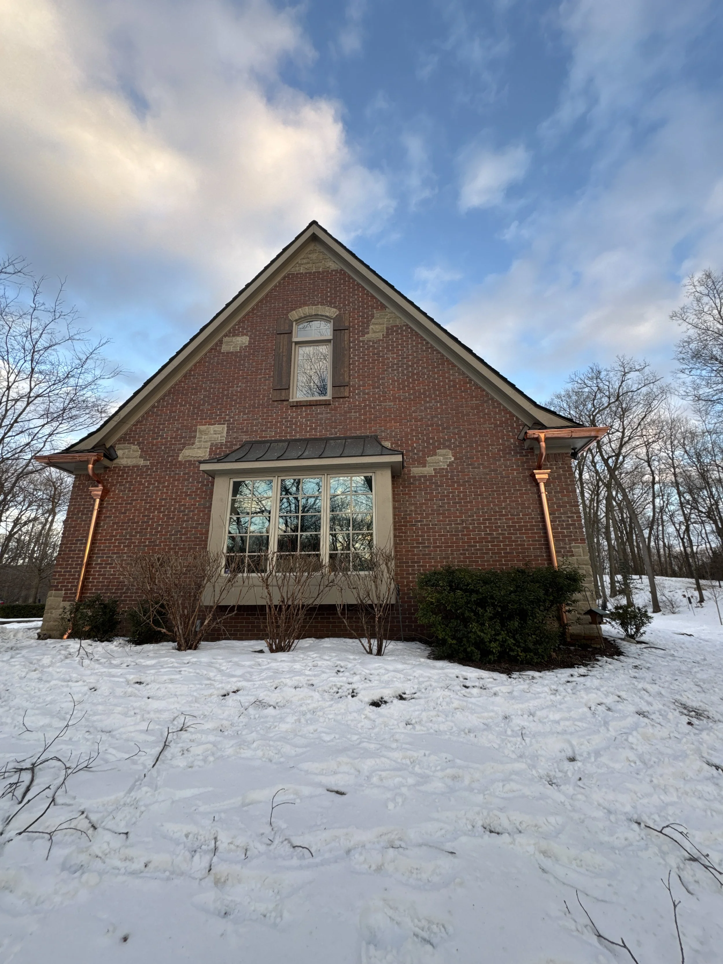 A brick house with a large bay window, small bushes in front, and a snow-covered yard under a partly cloudy sky. home in Milford installed seamless copper gutters with dads seamless gutters, radius gutter, copper gutter and copper pipe downspouts.