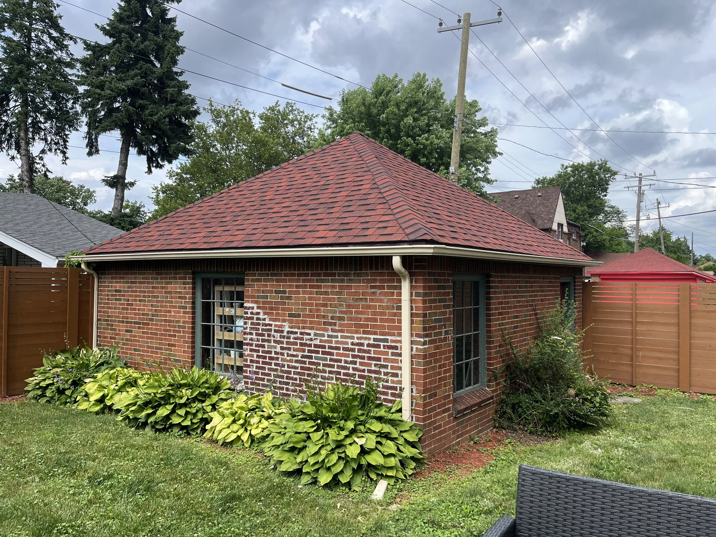 A small brick house with a red shingle roof, green-framed windows, surrounded by green bushes and a manicured lawn, with a wooden privacy fence and utility poles in the background.dads seamless gutters cleaned gutters