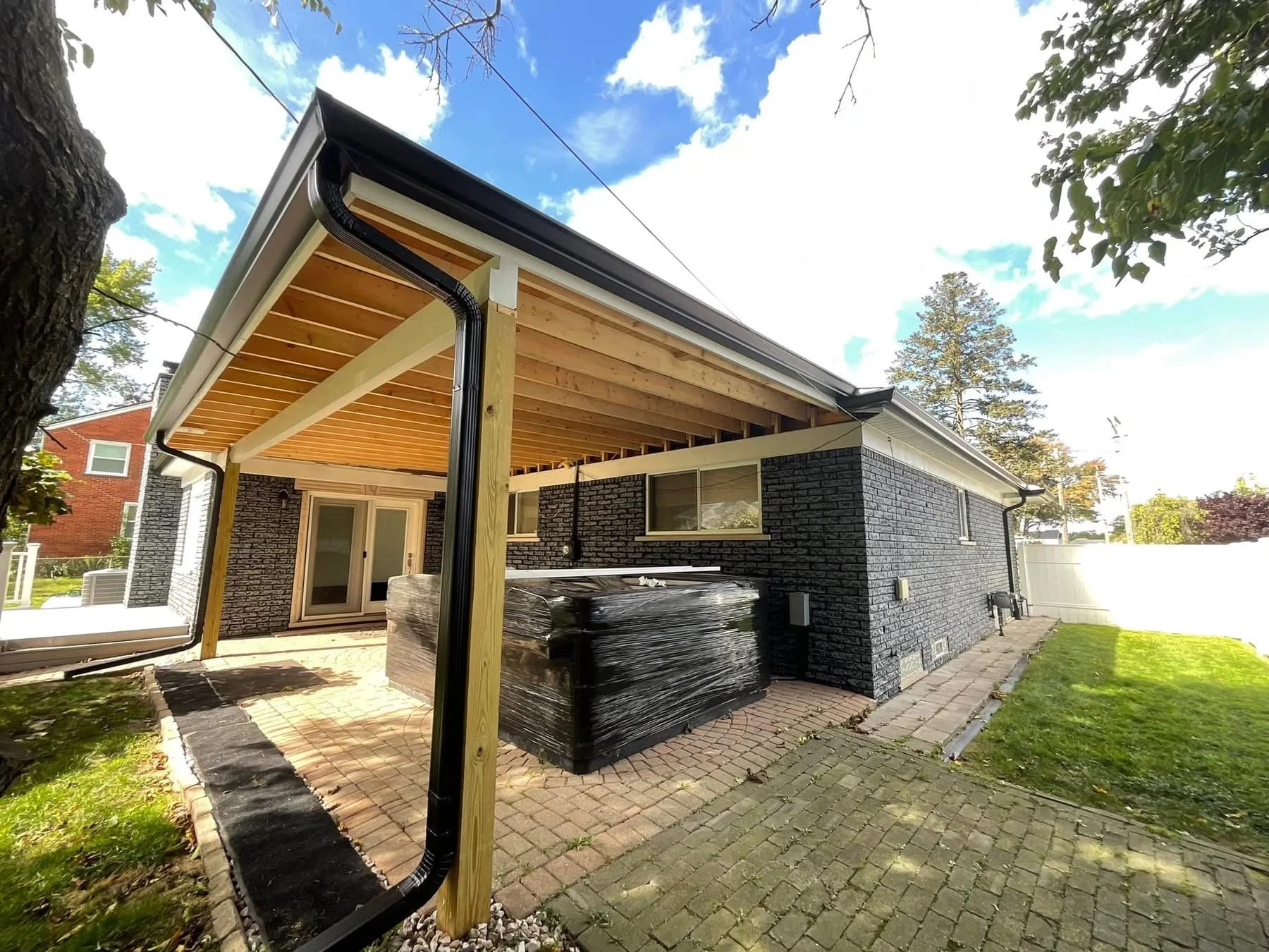 Backyard of a house with a patio, brick walkway, and a covered porch area under construction. The house has dark painted brick walls and white trim, and black seamless gutters installed by dads seamless gutters in michigan