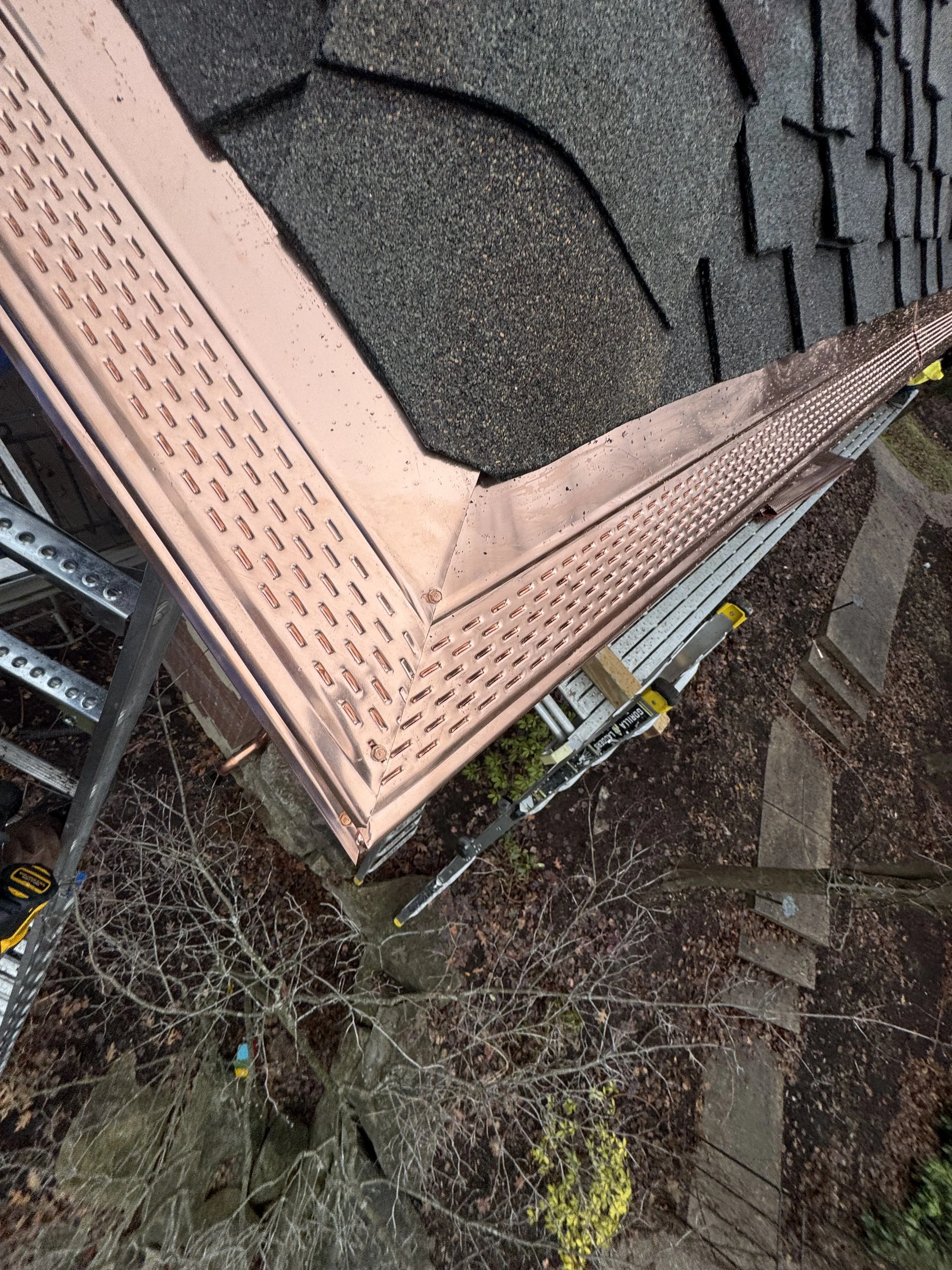 Close-up of a roof corner with asphalt shingles and copper gutters with copper leaf guards seen from above, with leafless trees and stepping stones in the background. dads seamless gutters