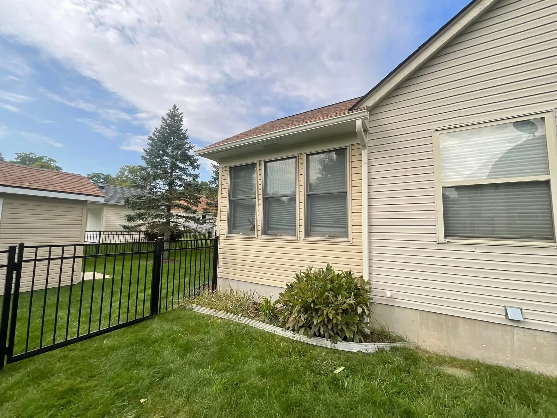 Backyard with green grass, shrubs, a black fence, and beige house with vinyl siding and several windows under a partly cloudy sky.dads seamless gutters installed clay seamless gutters in Michigan 
