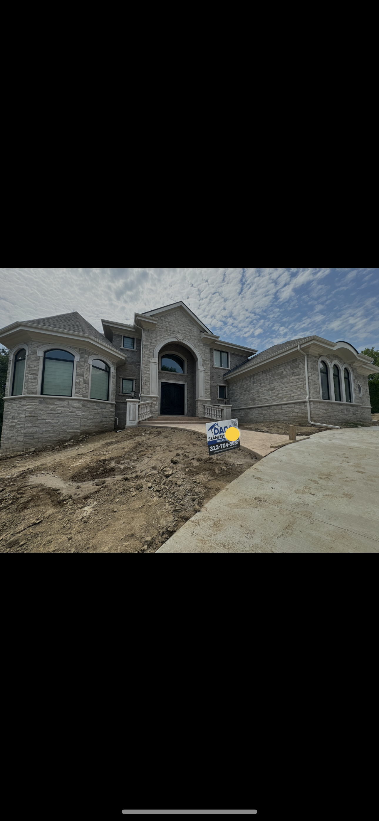 Newly constructed large stone house with arched windows and a front porch, surrounded by dirt and a concrete driveway under a partly cloudy sky. dads seamless gutters installed there new white gutters