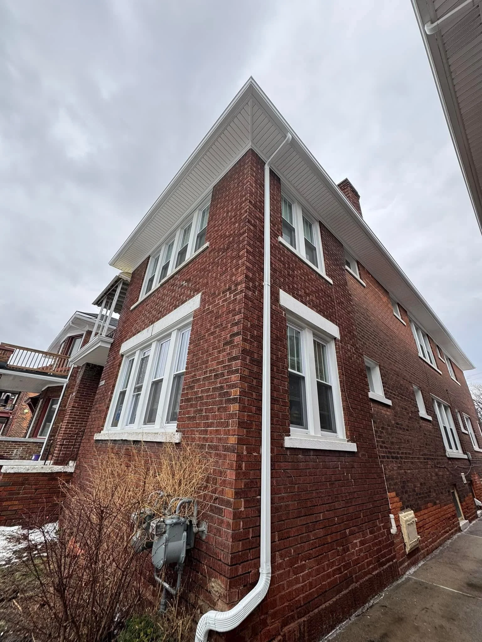 A photo of a corner of a brick house with multiple white-framed windows, white seamless 5' gutters and downspout, installed by dads seamless gutters on a duplex for the city of detroit michigan