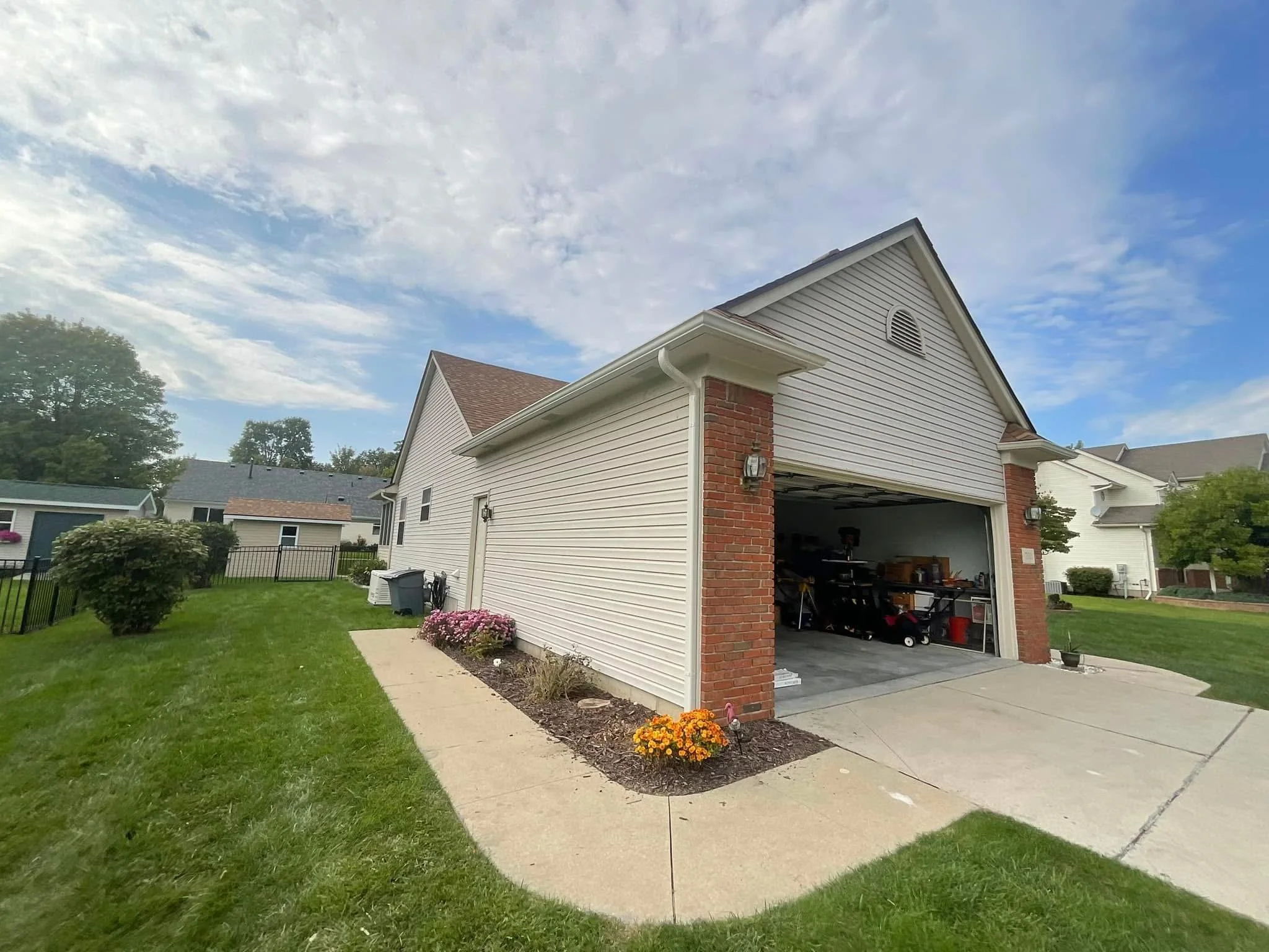 Front of a house with a one-car garage, partly open, surrounded by green lawn and small garden flowers, under a partly cloudy blue sky.new house new gutters installed in macomb Michigan 