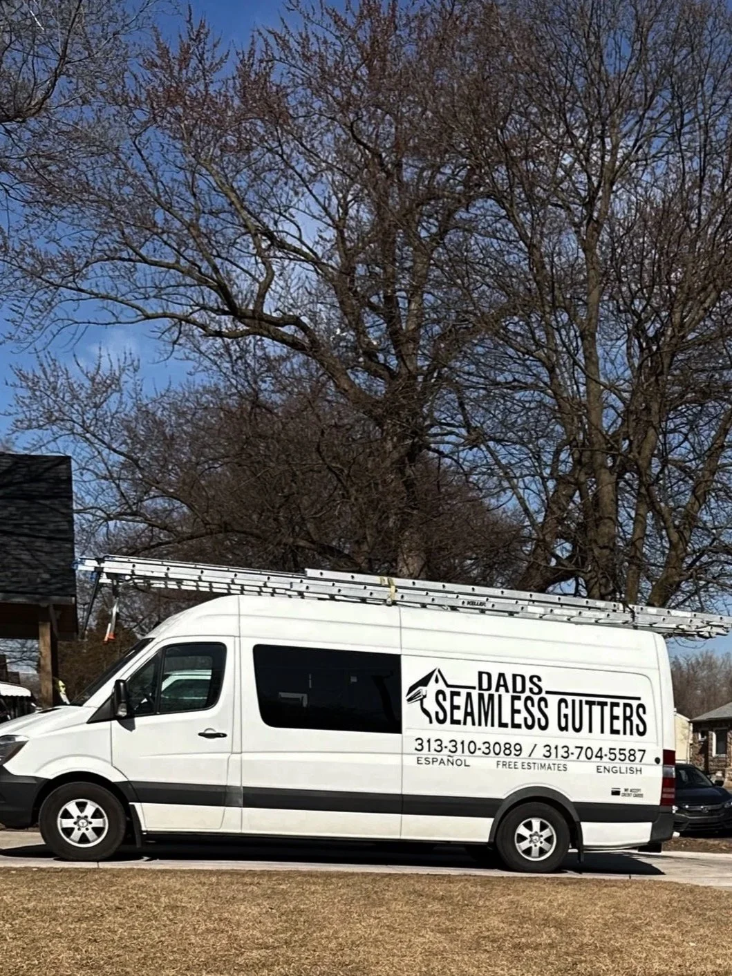 White service van with the logo Dads Seamless Gutters, ladder on top, parked on a residential street with leafless trees and houses in the background.