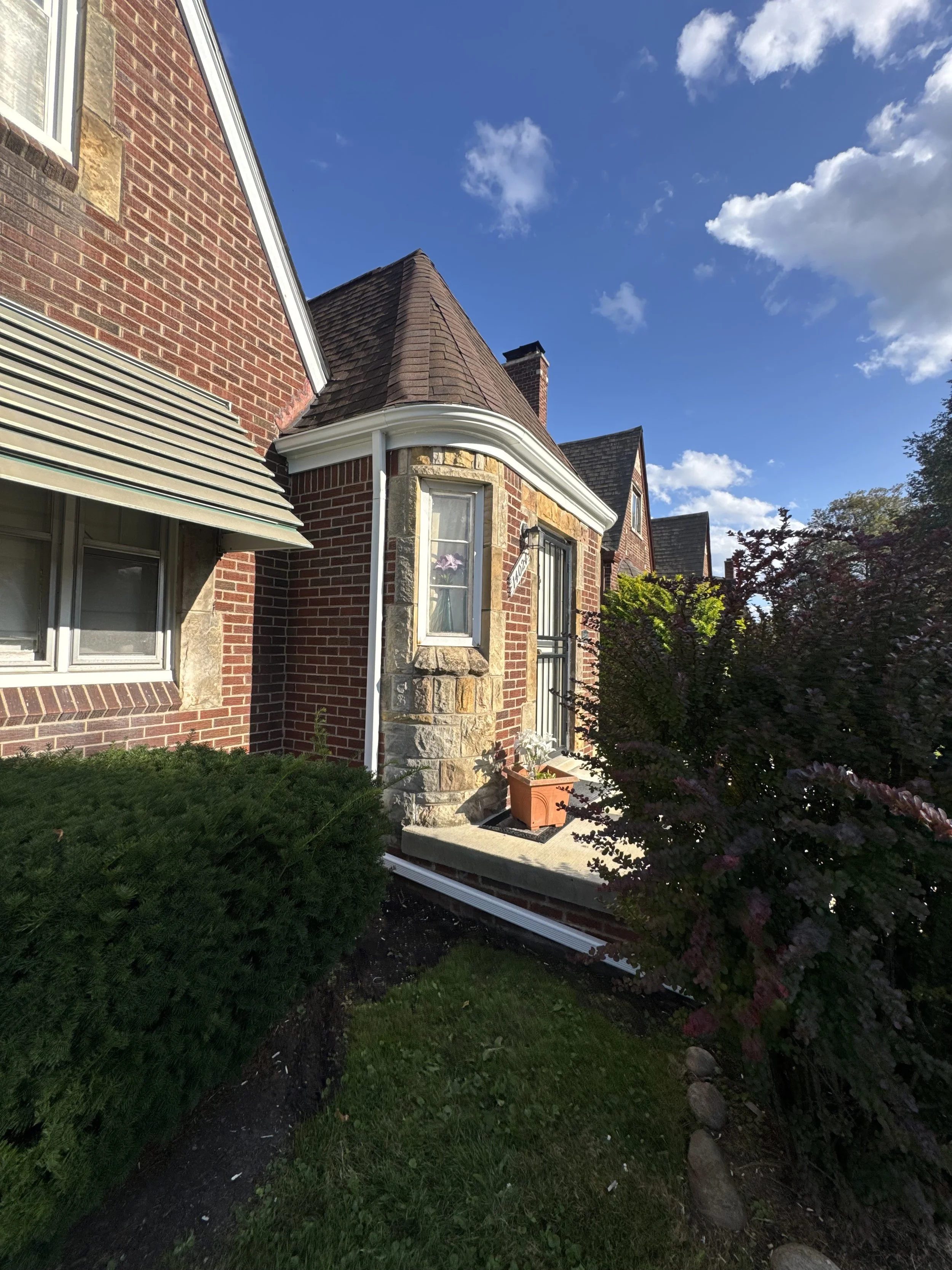 Side view of a brick house with a small porch, bay window, and decorative stonework, surrounded by greenery and a blue sky with scattered clouds. dads seamless gutters installed seamless gutters 5' radius gutter on front porch to secure clients porch