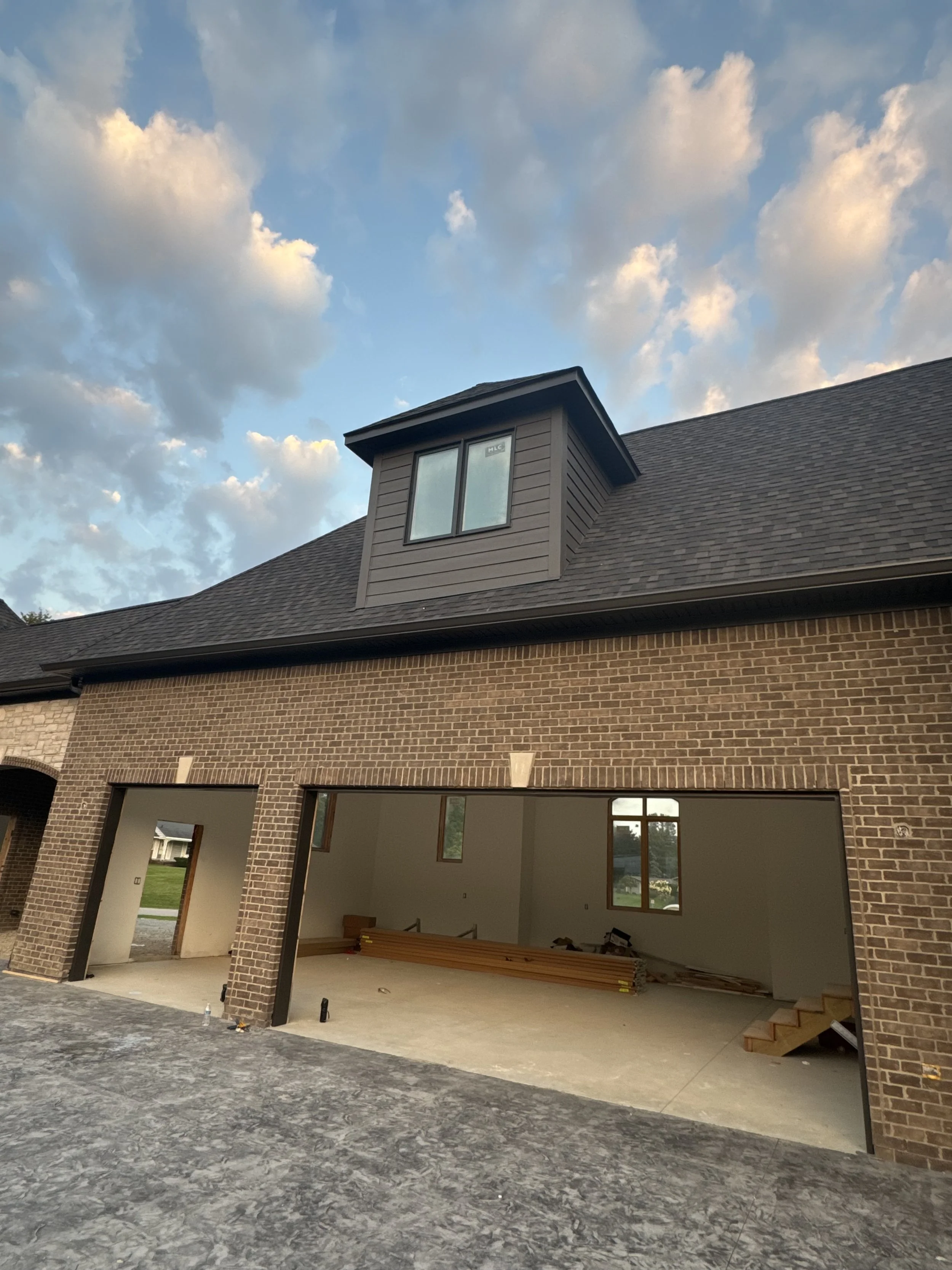 Photo of a house under construction with an open garage area, brick exterior, and a dormer window on the roof, under a cloudy sky. dads seamless gutters worked on this home installed black 6' gutters in northville michigan 