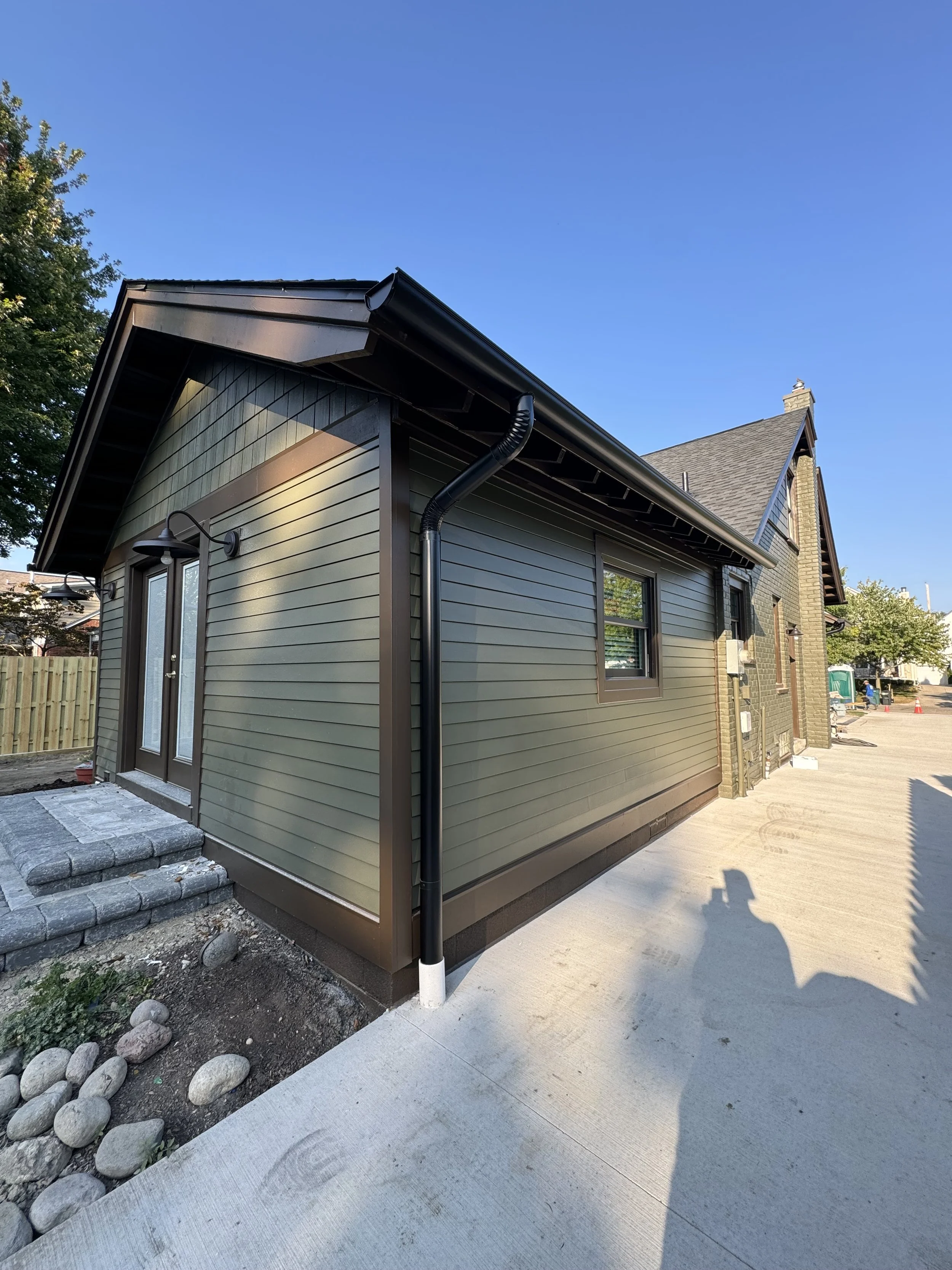 Side of a newly constructed house with green siding, a black gutter, and a concrete pathway, under clear blue sky. dads seamless gutters installed gutters black 6'.