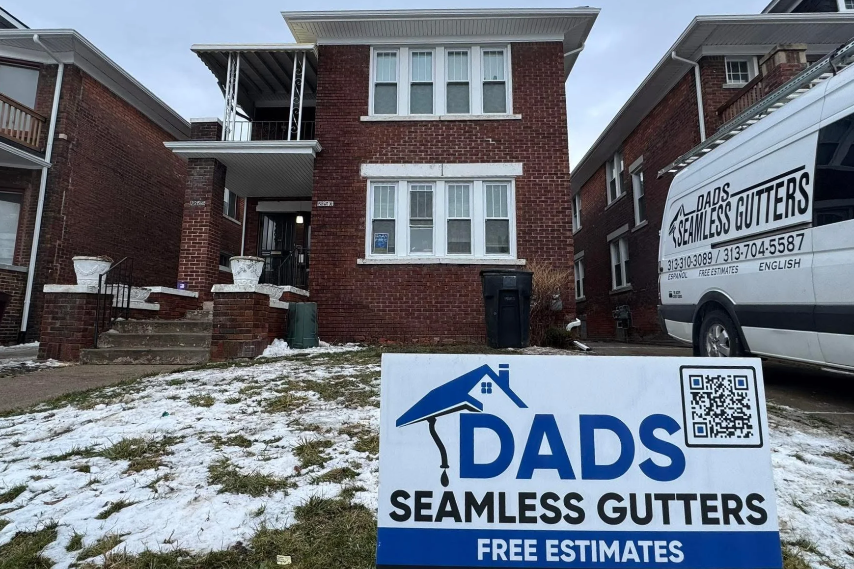 Front view of a brick residential building with a discolored white sign in the grassarea in front advertising Dads Seamless Gutters, offering free estimates. There is snow on the ground. working year round protecting home