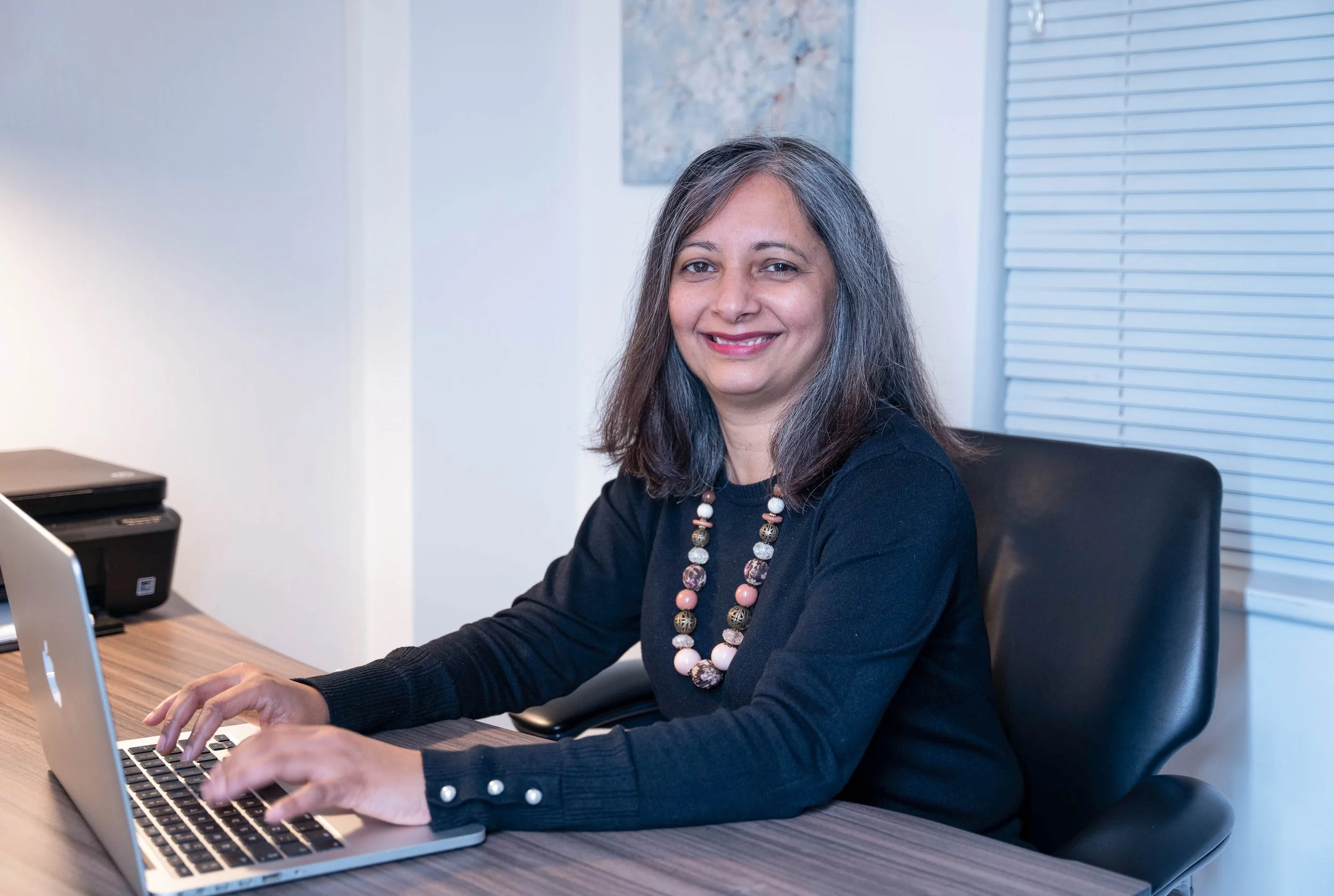 A woman with gray hair, wearing a black top and a colorful beaded necklace, sitting at a desk and working on a laptop in an office setting.