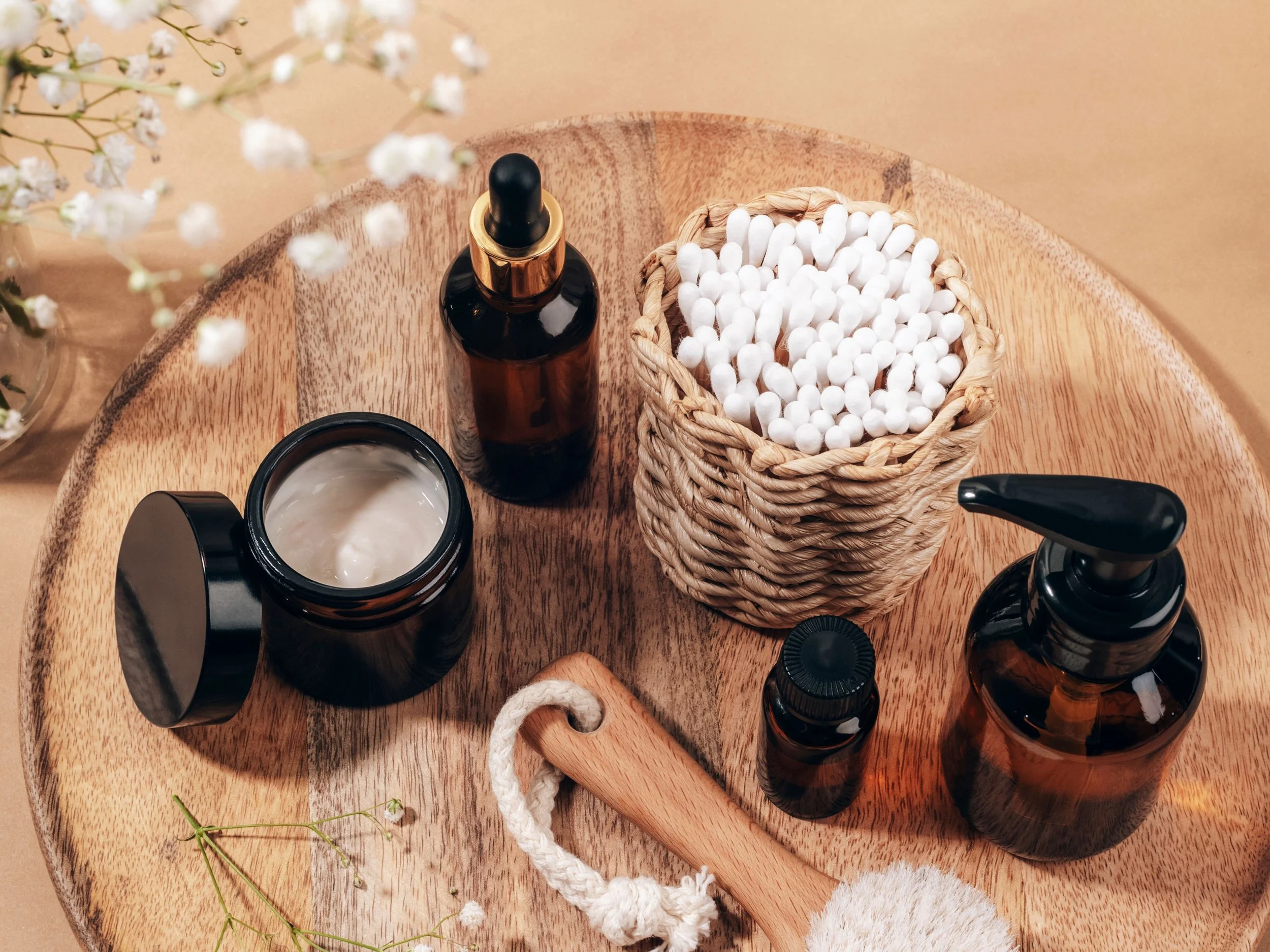 A wooden table with skincare products including a dark amber dropper bottle, a black spray bottle, an open black jar with cream, a small dark amber bottle, a woven basket filled with cotton swabs, and a wooden facial massager, with white flowers in the background.