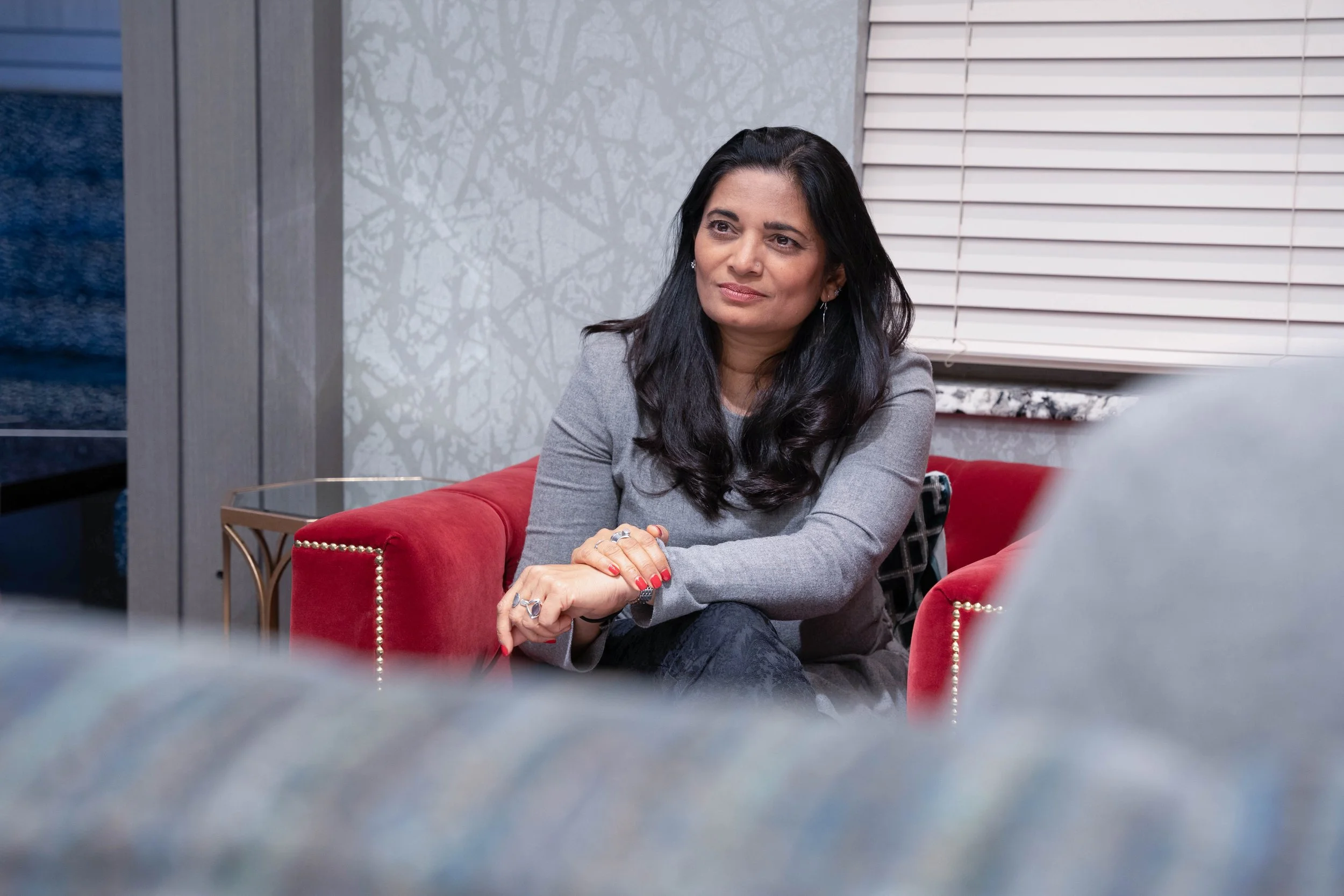 A woman sitting on a red velvet sofa in a room with patterned wallpaper and closed white blinds, appearing to be engaged in a conversation.