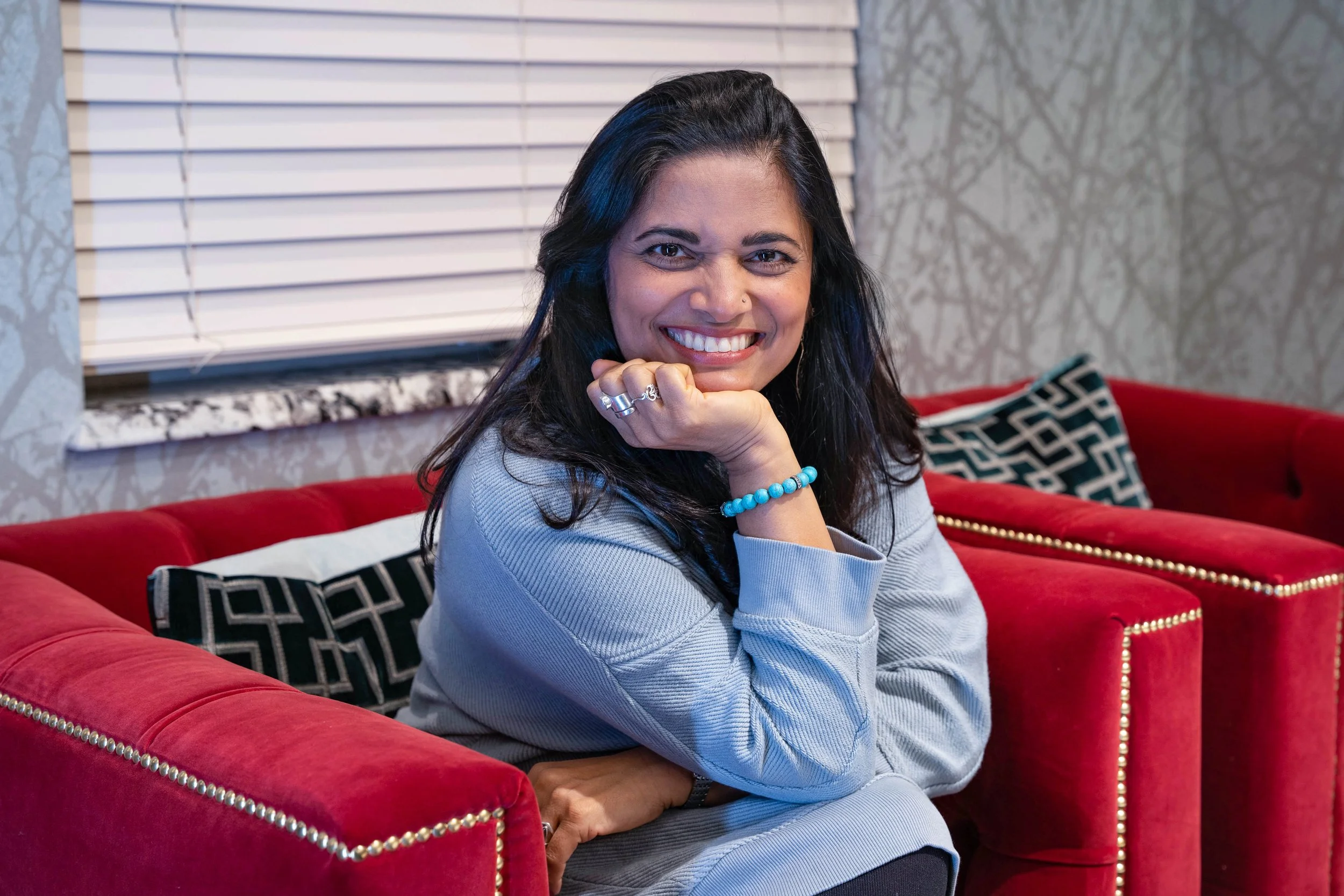 A woman with dark hair and a bright smile sitting on a red couch in a cozy room with patterned wallpaper and white window blinds.