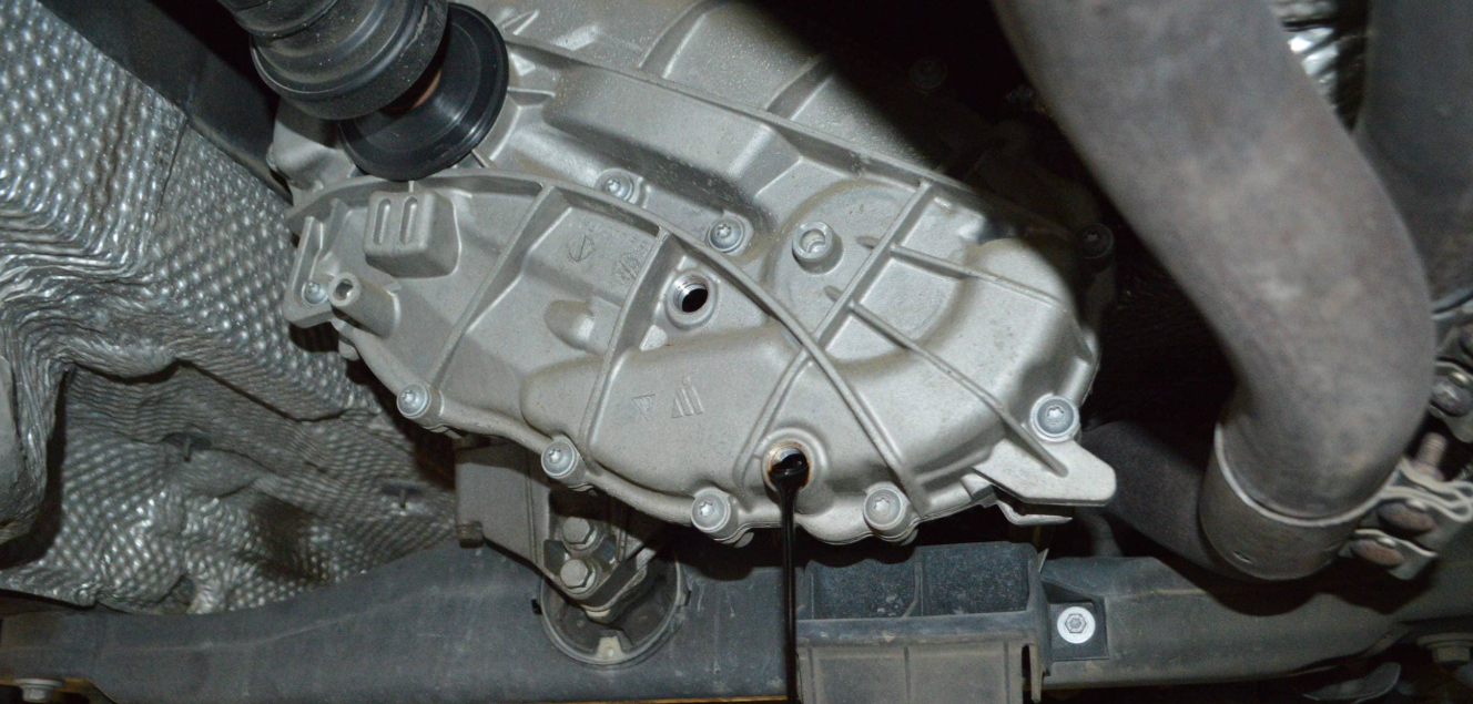 View under a vehicle showing a transmission with oil leaking from the drain plug, and surrounding components including heat shielding and hoses.