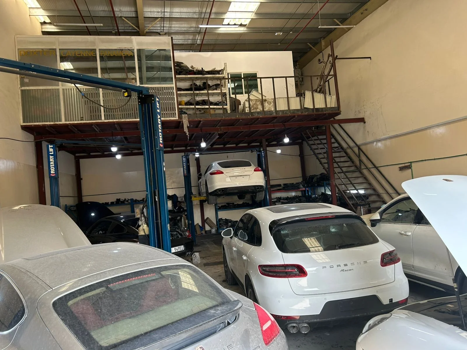 Several luxury cars, including Porsche Macan and Porsche Cayenne, parked inside a multi-level car repair garage with a hydraulic lift, a mezzanine level with shelves and equipment, and overhead lighting.