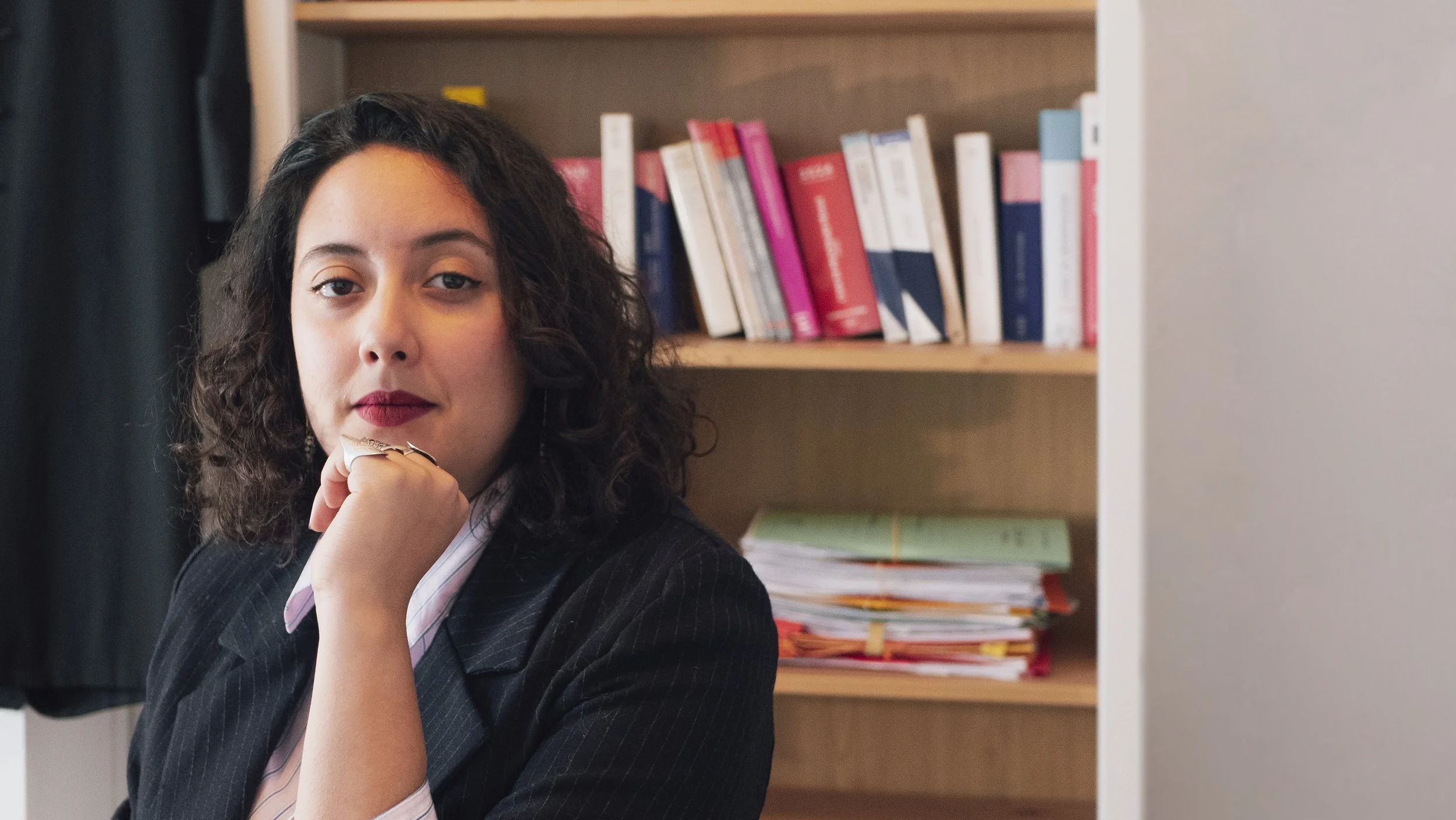 Femme avec cheveux bouclés, portant un costume noir, assise dans un bureau, avec une bibliothèque en arrière-plan.