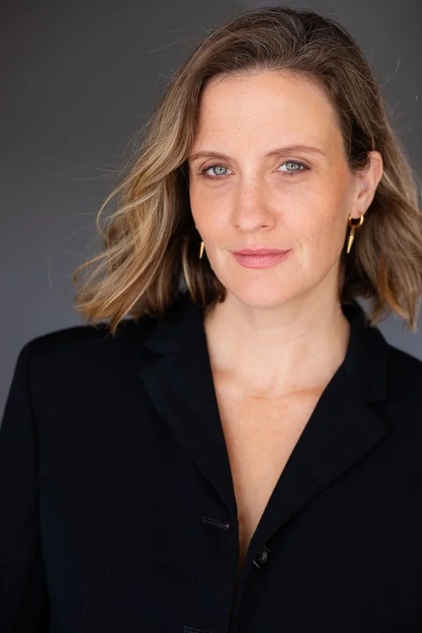 Headshot of performer and mother with shoulder-length wavy blonde hair, wearing a black blazer and gold earrings, against a dark gray background.