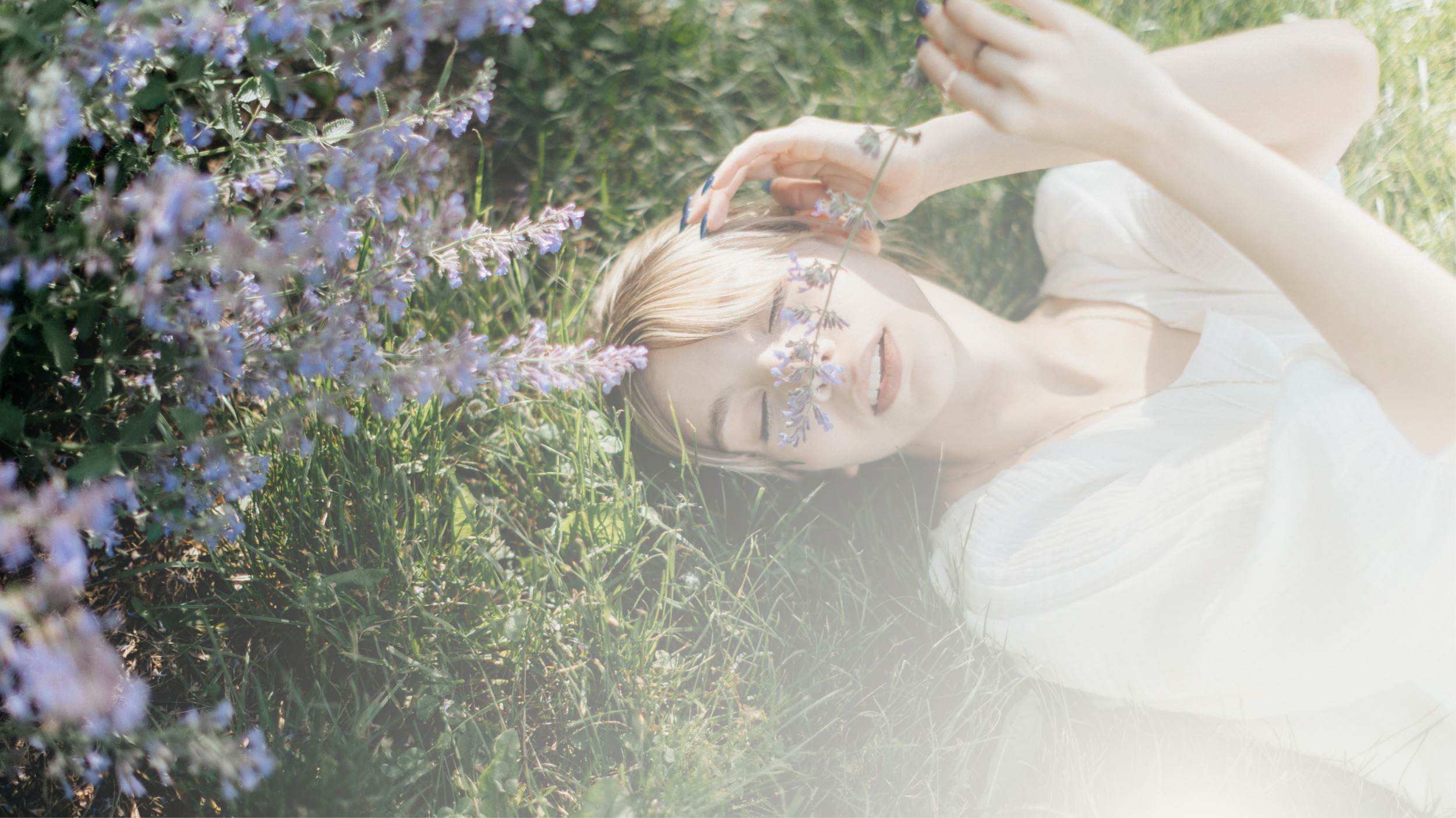 A woman lying on the grass outdoors among purple flowers, with eyes closed, touching her forehead with one hand, and a peaceful expression.