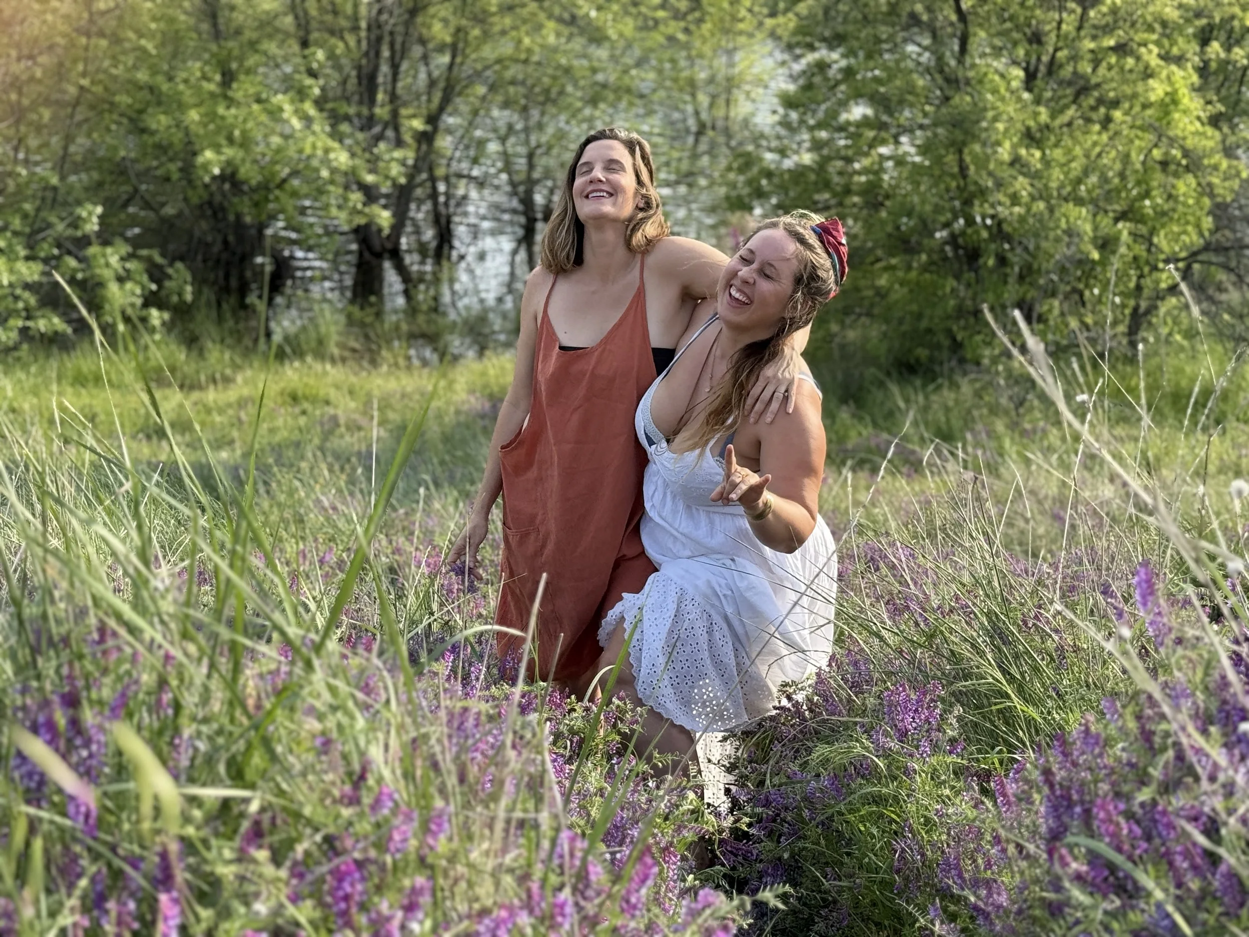 The woman of the musical group Wildflower in a grassy field with purple flowers and green trees in the background, smiling and having fun together.