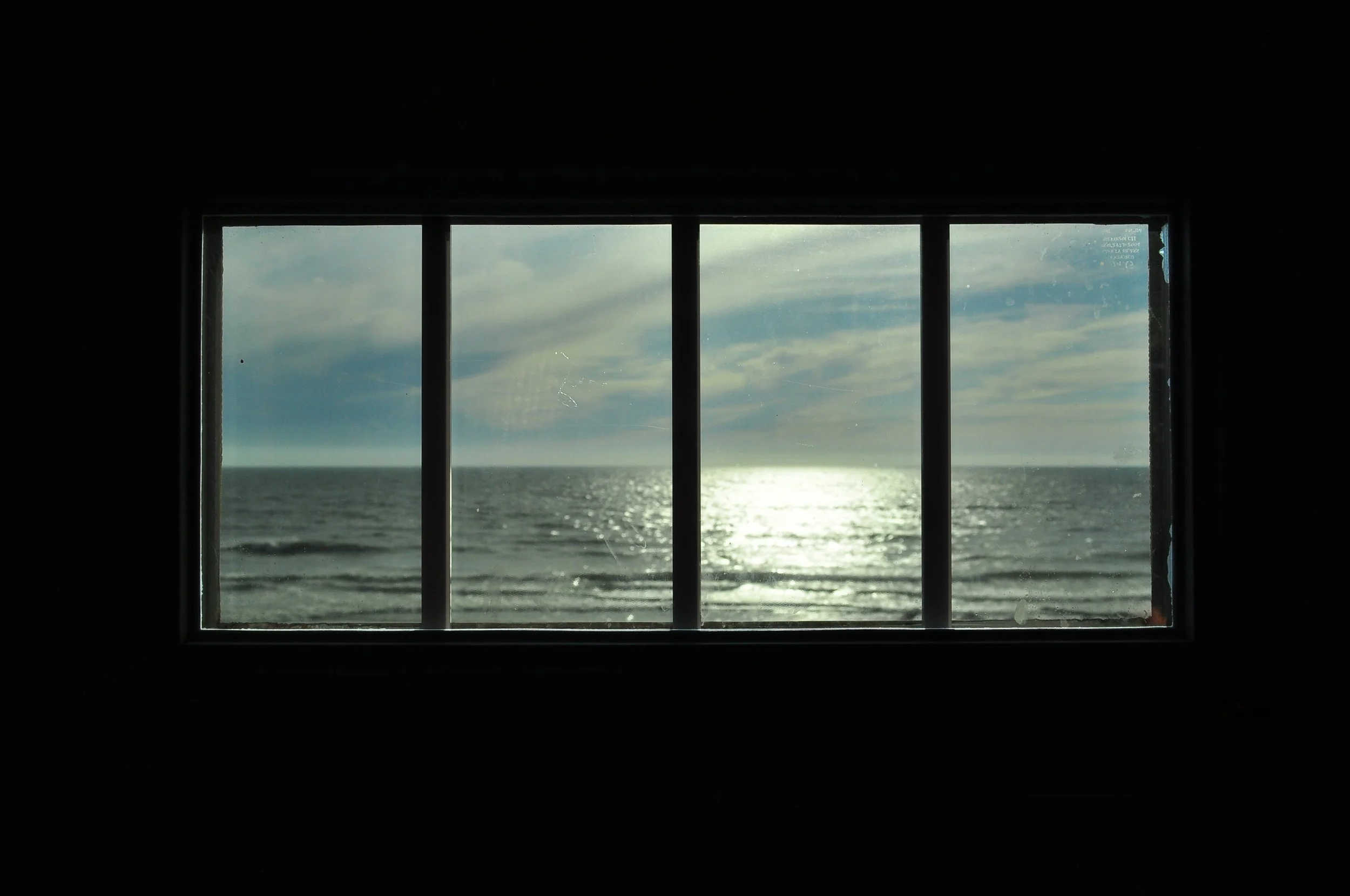 View of a seascape through the front door window of a skoolie, showing the ocean, sky, and sunlight reflecting on the water.