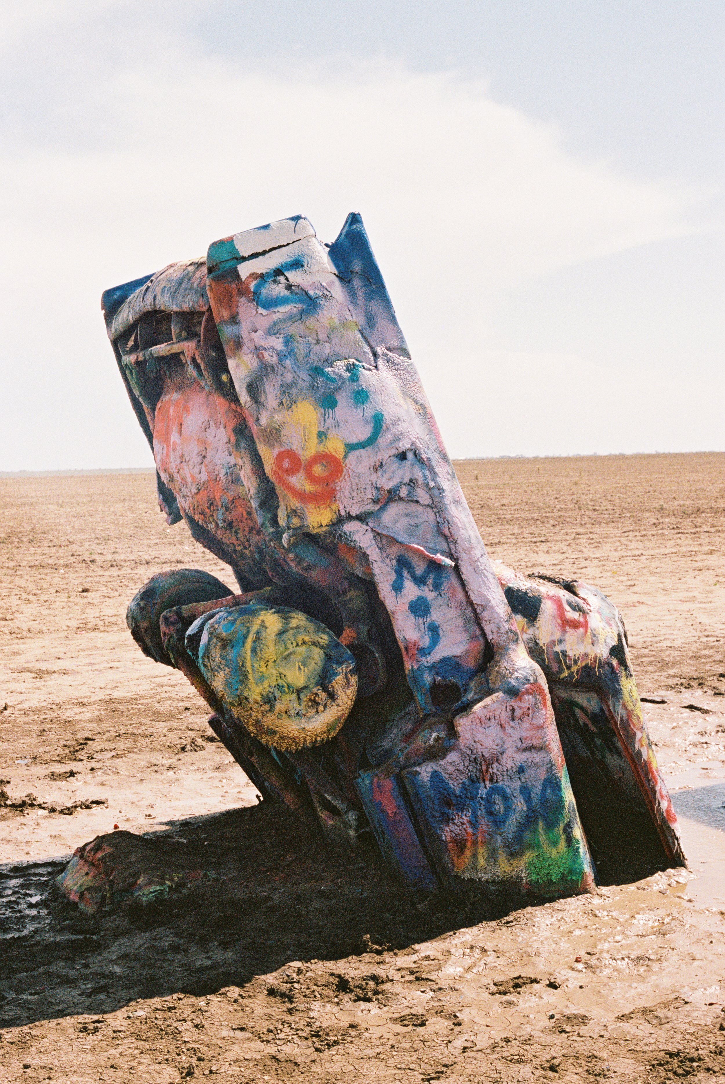 A colorful, spray-painted Cadillac, partially buried in the ground in a barren, dry landscape under a cloudy sky at Cadillac Ranch. 35mm film.