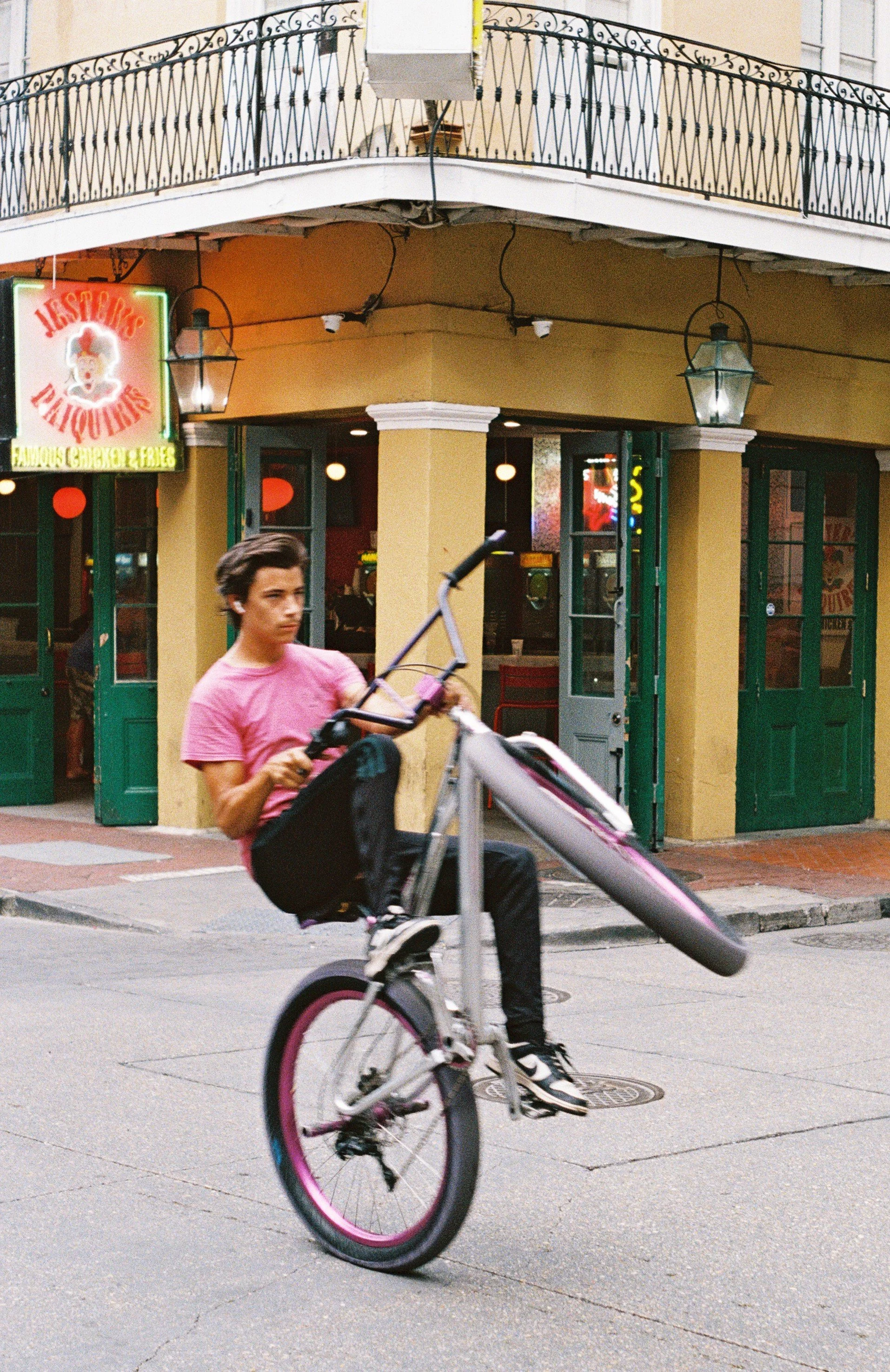 Young man performing a wheelie on a bicycle in front of a colorful restaurant with a yellow exterior and green doors.
