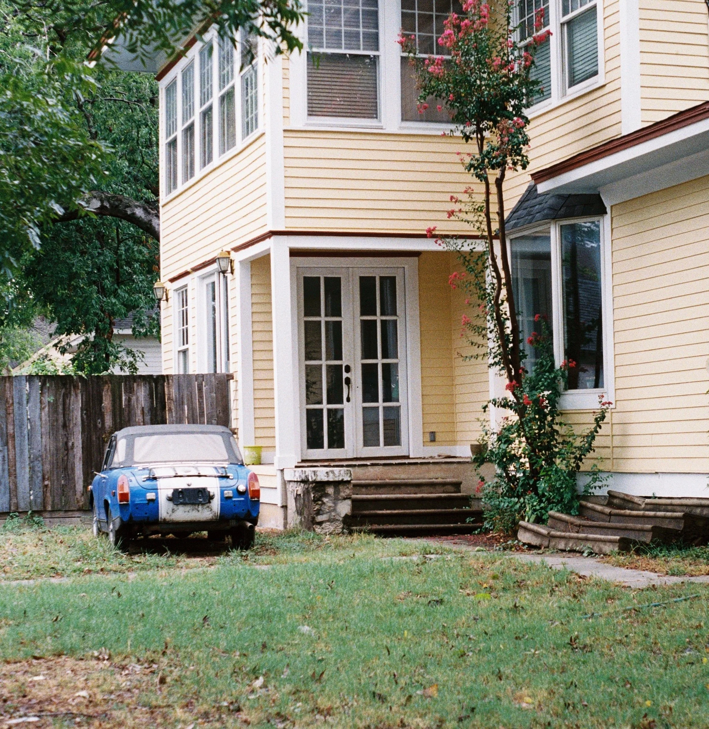A yellow, two-story house with white trim and multiple windows, a small front porch with a few steps, and yards with grass and a tree with pink flowers. An old, blue car with a partially worn exterior is parked close to the house, Denton