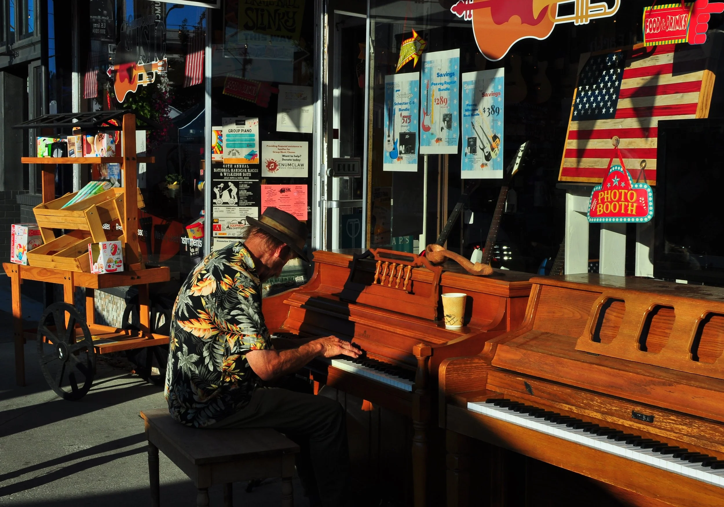 An older man wearing a colorful Hawaiian shirt and a wide-brimmed hat, sitting outdoors and playing a piano. Behind him is a storefront with posters, a guitar, and an American flag sign that says 'Photo Booth'. Enumclaw, Washington