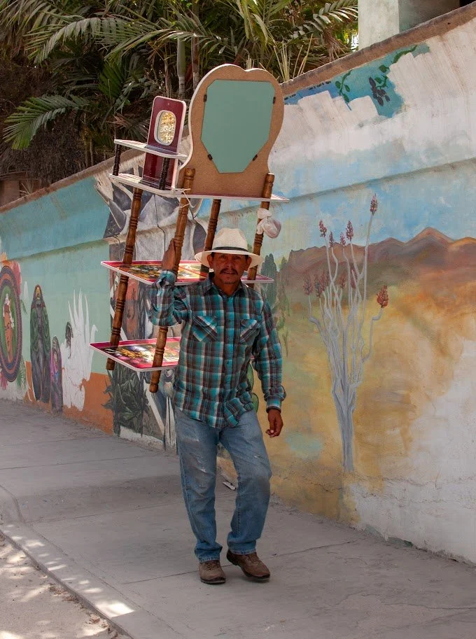 Man in plaid shirt and straw hat walking on sidewalk, balancing a chair on his shoulder, with colorful mural of trees and mountains on wall behind him. Baja Mexico