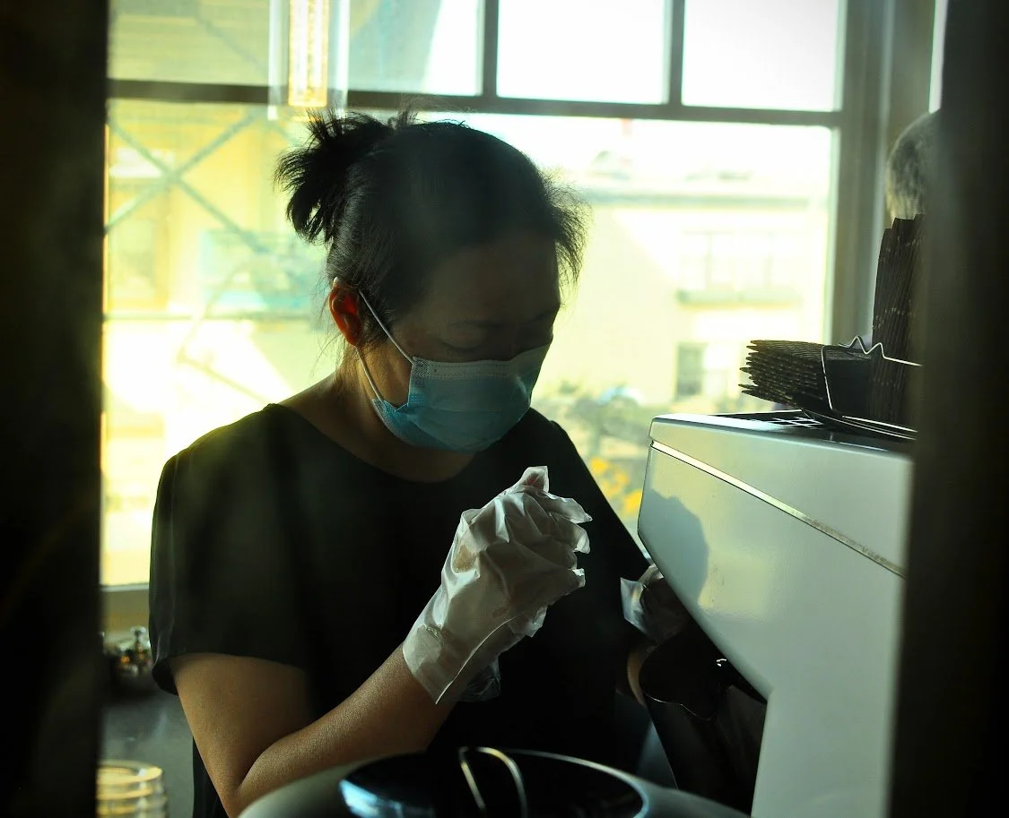 A woman wearing a face mask and gloves making coffee with a large window in the background letting in natural light. Seattle Washington